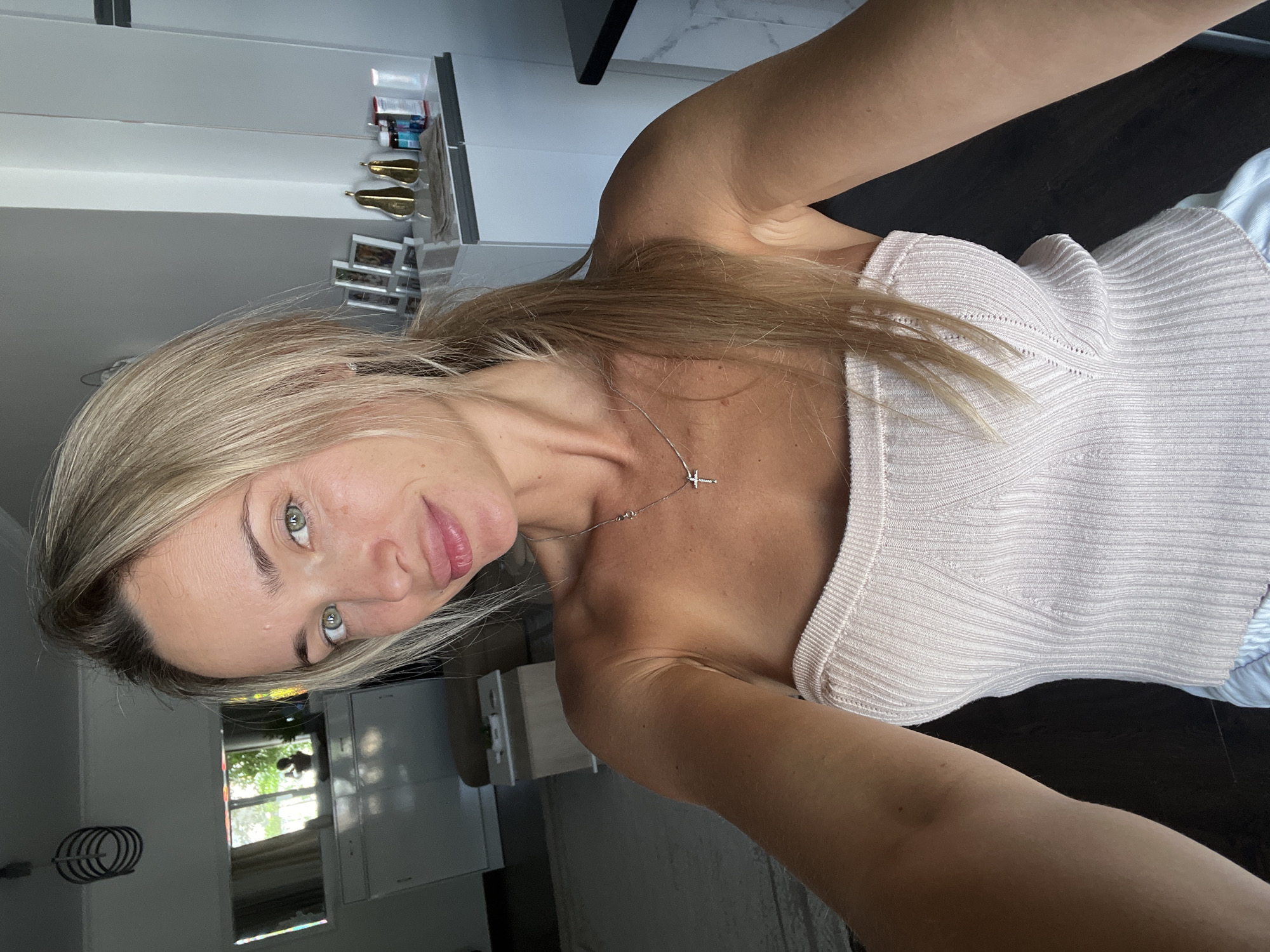 A young, blonde woman with green eyes takes a selfie in what appears to be a bright, modern apartment with natural light. She is wearing a light pink strapless top and a silver cross necklace. Her hair is pulled back in a ponytail. In the background, there is a mirror reflecting a window with greenery, a white dresser, and a shelf with decorative items. The overall impression is one of a casual, indoor setting. The location is not identifiable as Sharm El Sheikh, Egypt.