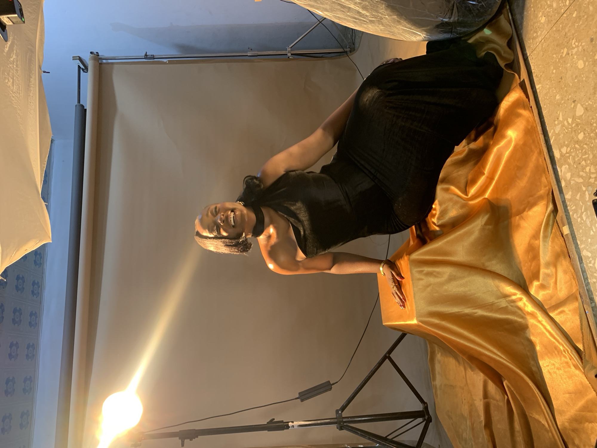 The image depicts a woman in a black, one-shoulder dress posing in a studio setting. She is seated on a golden fabric draped over a cubic platform. The woman is smiling and looking to her left. A bright light source is visible to the left of the frame, illuminating the scene. In the background, a tan backdrop is visible, along with studio equipment like a light stand. The floor appears to be tiled. The overall impression is a professional photoshoot. The location of Gura Top, Nigeria is not evident from the visual information in the image.