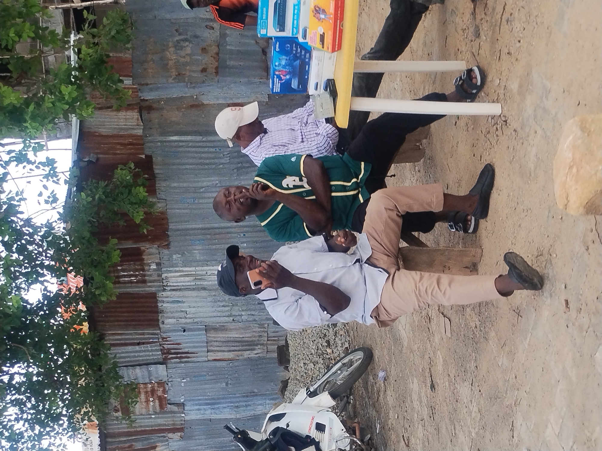 The image depicts an outdoor scene, possibly in Jos, Nigeria. In the foreground, three men are seated. The man on the left is wearing a light-colored collared shirt, tan pants, and black shoes, and is holding a phone to his ear. He is seated on a wooden stool. To his right, a man in a green and white baseball jersey with arms crossed is also seated, with his index finger to his lips. Further to the right, a man in a checkered shirt and a white cap is seated, looking down, with his hands near a yellow table. The table holds several electronic devices in boxes. The background consists of a corrugated metal wall, with some trees and foliage overhead. A white motorcycle is parked to the left of the frame. The ground appears to be dirt and gravel.