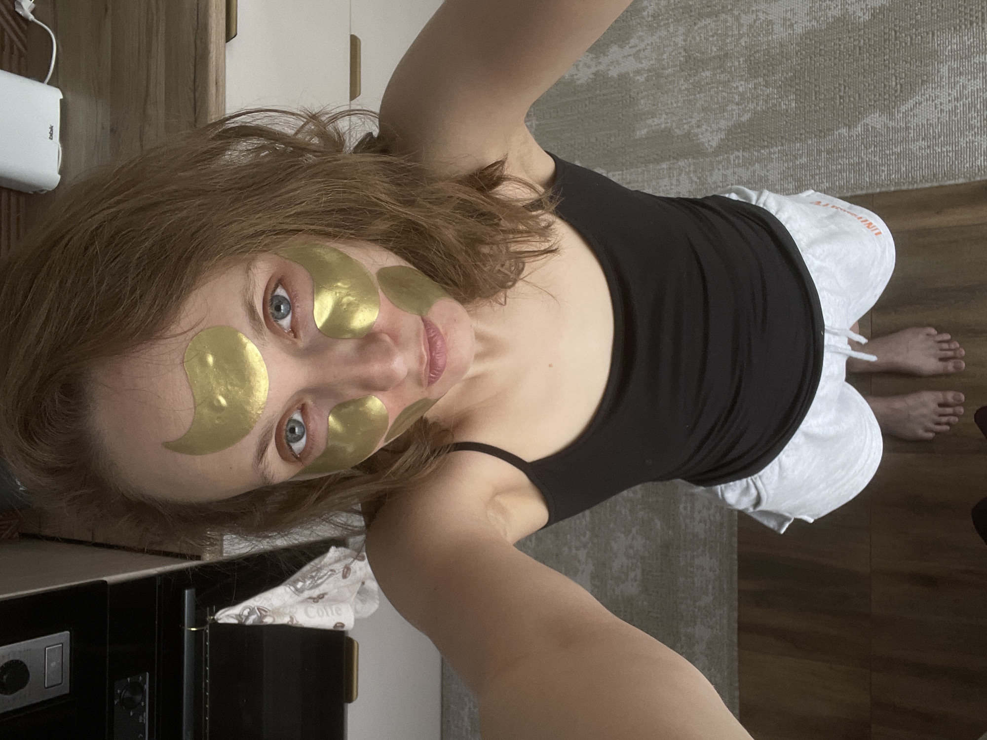 A fair-skinned woman with light brown hair poses in an overhead shot. She wears a black tank top and white shorts. Gold facial patches are adhered to her forehead and under her eyes. Her bare feet are visible in the lower right corner of the frame. The background consists of a mottled gray rug, dark wood flooring, and various pieces of furniture including a white electronic device and a black cabinet with silver dials. The location is Unknown, Russia.