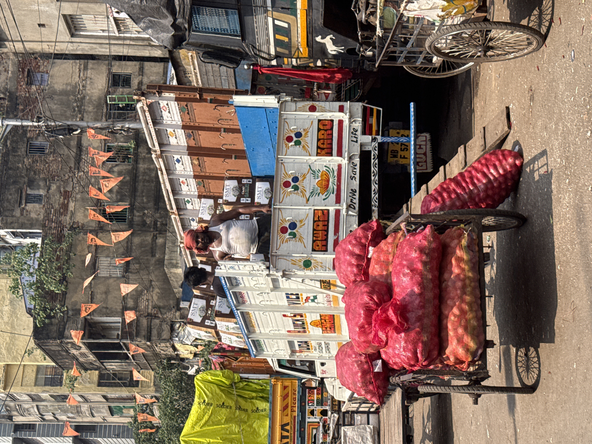 Here is a detailed description of the image:
**Scene Overview:** The image depicts a street scene in Kolkata, India, showing a man standing on the back of a brightly decorated truck, with a cycle rickshaw loaded with sacks of onions in the foreground. Buildings line the street, decorated with rows of orange pennants.
**People:**
* A man wearing a white tank top and a red headscarf is standing on the back of the truck. He appears to be looking at something or someone.
* A second man is partially visible on the truck, wearing a dark-colored shirt.
**Subjects & Objects:**
* **Truck:** The truck is painted with colorful designs and words, including "KARO" and "Drive Save Life." Various logos and smaller text are also visible.
* **Cycle Rickshaw:** A three-wheeled cycle rickshaw is positioned in the foreground.
* **Onions:** The rickshaw is heavily loaded with multiple large sacks of red onions.
* **Buildings:** Buildings line the street, showing signs of age and wear.
* **Orange Pennants:** Rows of orange pennants hang from the buildings, adding a decorative element to the street scene.
* **Traffic:** Additional vehicles, including trucks and possibly a car, are visible further down the street.
**Actions & Interactions:**
* The man on the truck is the primary subject of focus, but his specific action is unclear.
* The rickshaw is stationary, presumably waiting to transport or sell the onions.
* The environment suggests a busy street scene with commercial activity.
**Environment:**
* **Location:** Kolkata, India, as indicated by the vehicle registration plate and the general street environment.
* **Atmosphere:** The scene is filled with everyday urban life, showing a blend of commerce, transportation, and local culture.
* **Lighting:** The scene is well-lit, suggesting a daytime setting.