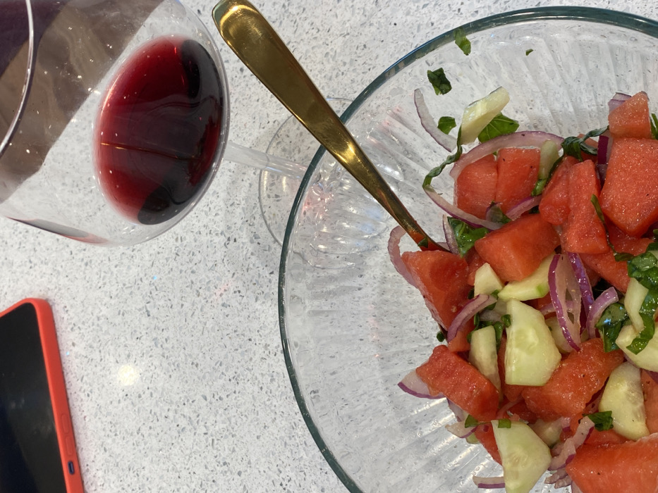 This image, taken in Ridgefield, United States, depicts a close-up, overhead view of a meal setup on a speckled, light-colored countertop. The primary subjects include a clear glass bowl containing a salad, a clear wine glass with red liquid, and a portion of a smartphone.
The salad bowl, occupying the lower and right-center portion of the frame, is made of clear glass with a vertical ribbed texture. It holds a mixture of diced red watermelon, sliced pale green cucumbers, and thin rings of purple-red onion, interspersed with small green leaf fragments, likely herbs. A golden-colored metallic spoon is partially submerged within the salad, positioned diagonally from the upper right toward the lower left of the bowl.
To the right of the bowl, a clear stemmed wine glass contains a dark red liquid, consistent with red wine. Its bowl is wide, and the liquid level is below its widest point.
In the upper left quadrant, partially visible, is a black smartphone with a bright red protective case. It lies flat on the countertop, its screen facing downwards or off. The countertop itself is a light gray or white surface with numerous small, dark speckles, suggesting a granite or quartz material. No individuals are visible in the frame. The scene suggests a prepared meal or beverage setting.