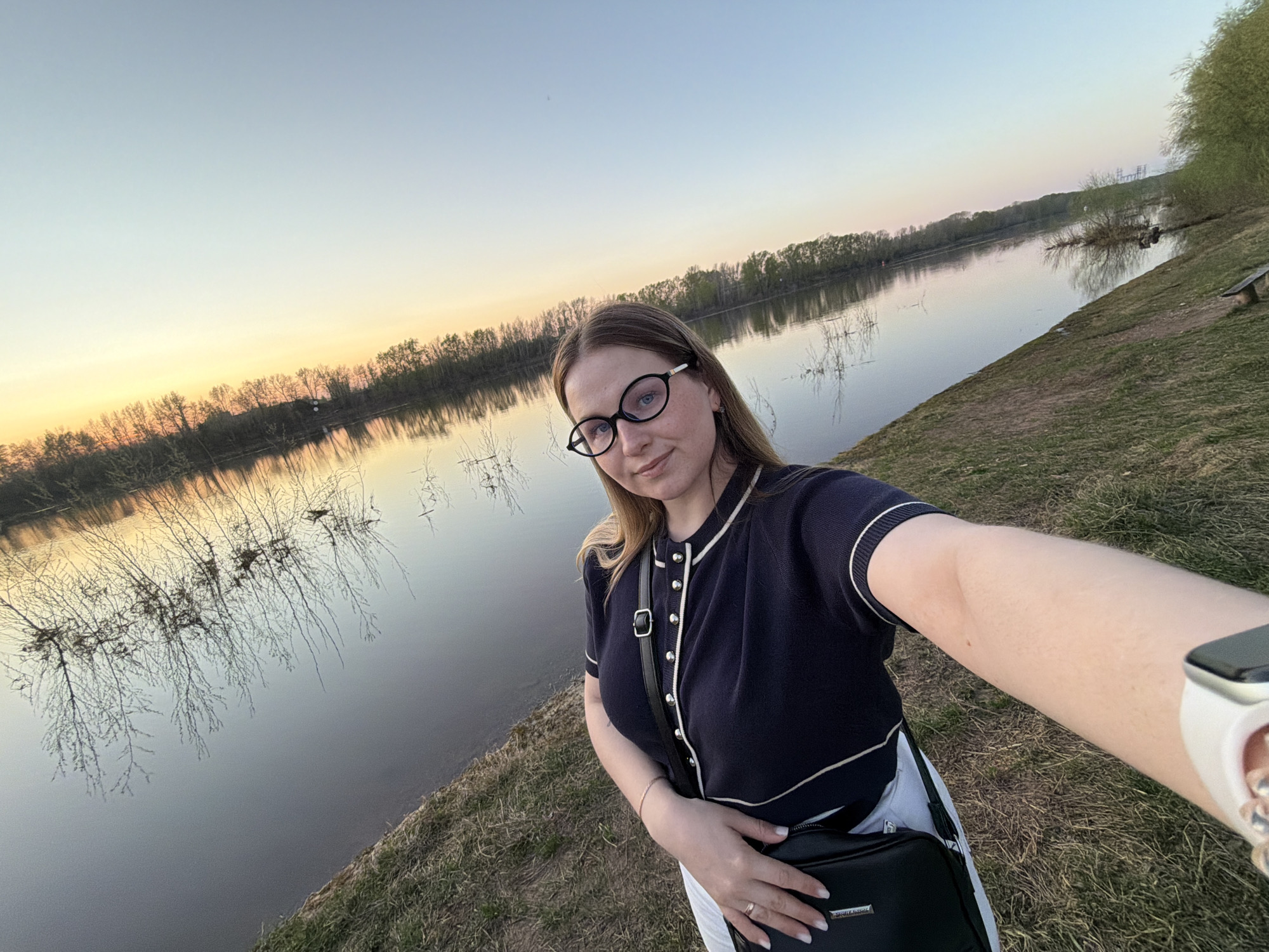 A woman in eyeglasses stands next to a river. She wears a dark blue, short-sleeved, button-down sweater with white trim and white pants. She also wears a black purse across her chest. The river is reflecting trees on its banks, and the sky is light blue and yellow. The location is Chaikovsky, Russia.