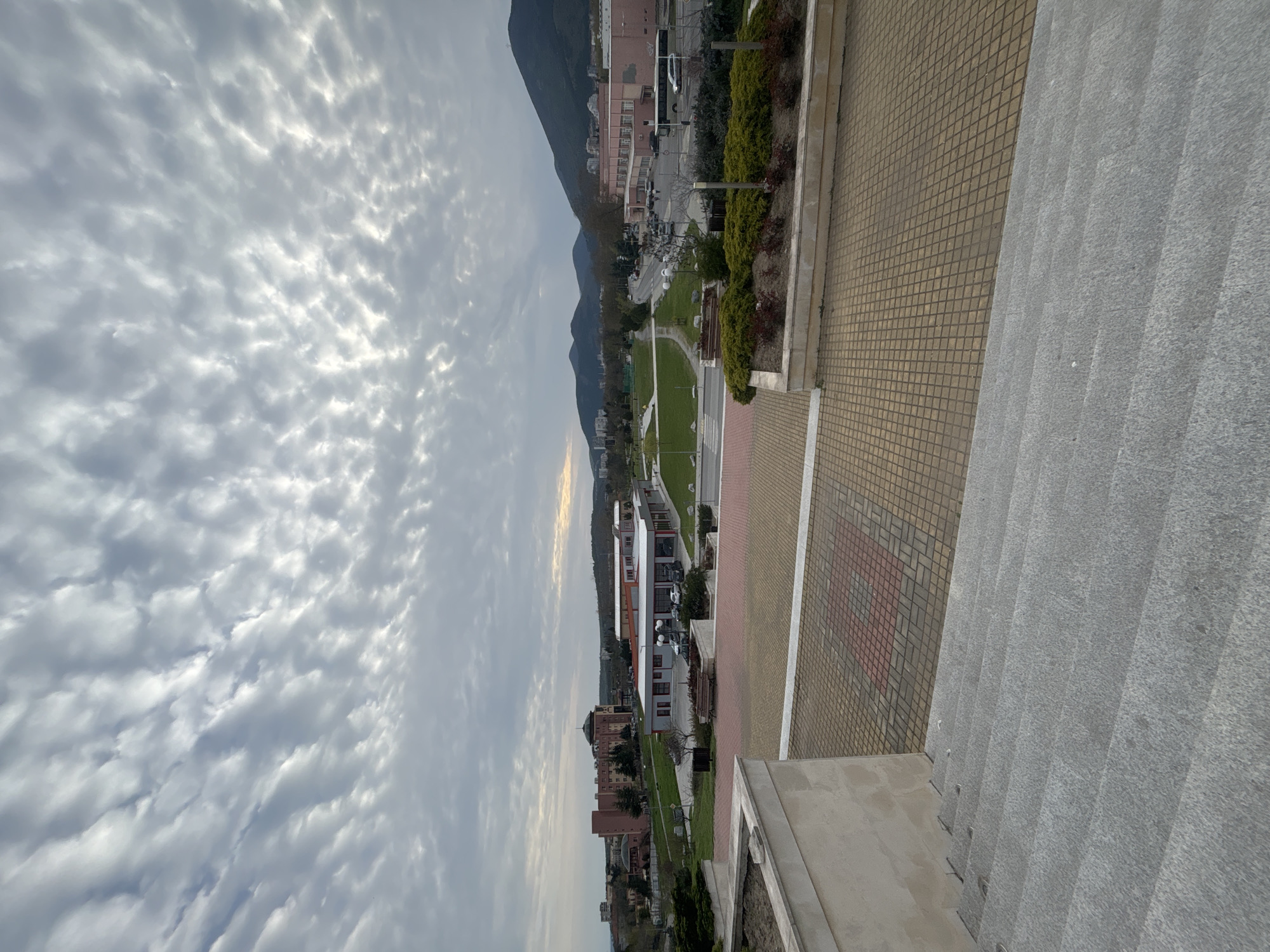 The image depicts a cityscape under a sky filled with altocumulus clouds. A mountain looms in the background. In the middle ground, buildings and a park are visible. In the foreground, there are steps and tiled pavement. The location is Unknown, Unknown. There are no people visible.