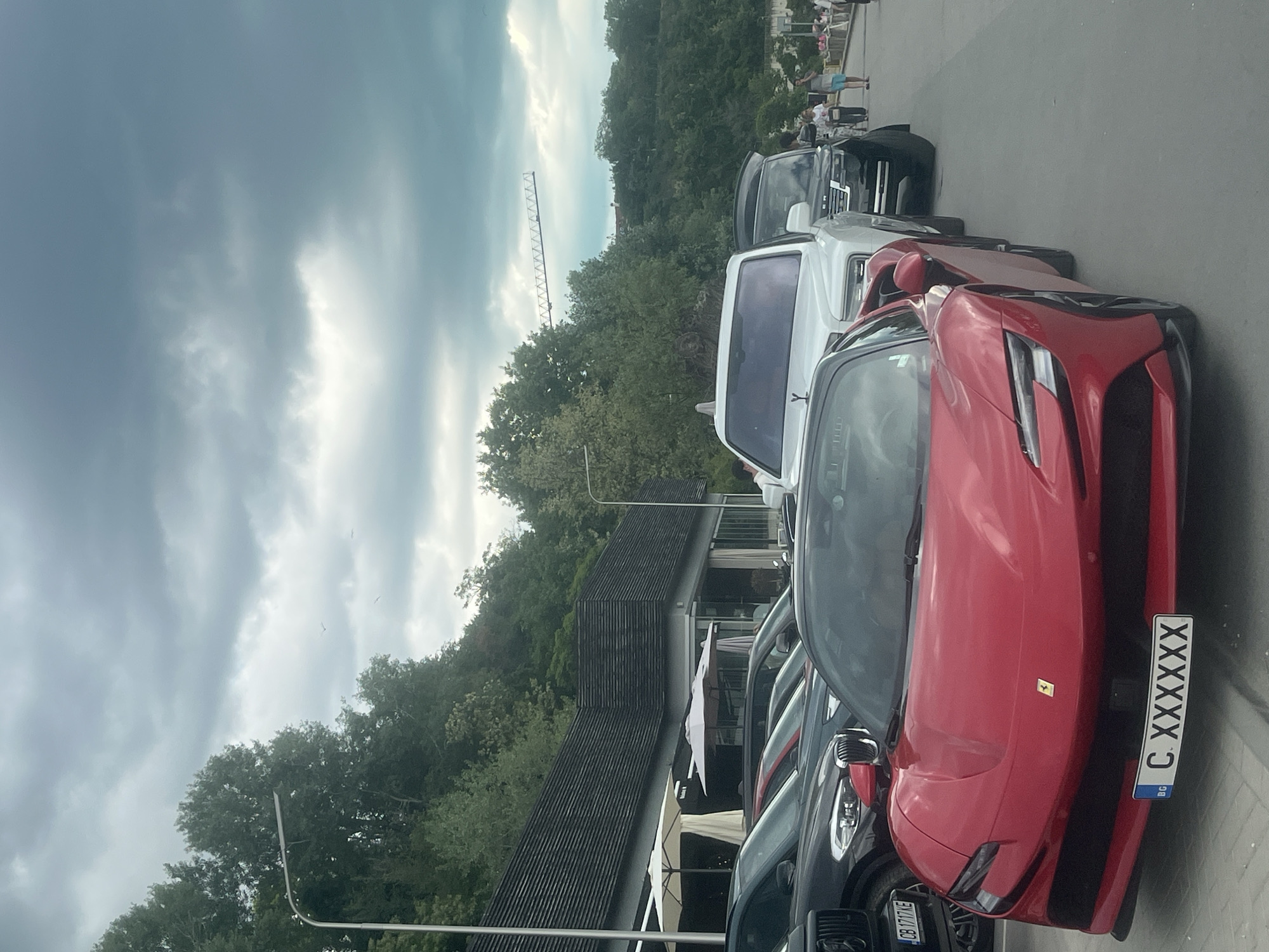 The image depicts a row of luxury vehicles parked along a street. Prominently featured is a red Ferrari with a Bulgarian license plate that reads "C XXXXXX." Behind the Ferrari is a white SUV, and further back, a black vehicle is visible. The setting appears to be an upscale location, possibly near a restaurant or establishment with outdoor seating, indicated by the awnings and a portion of a building roof in the background. In the distance, a construction crane is visible against a cloudy sky, suggesting an urban environment. The photo captures a scene with a concentration of high-end cars in what could be a wealthy area. This is a row of cars along a street near what seems to be a restaurant area with some greenery and construction visible in the background. In the top right corner, some people are visible.