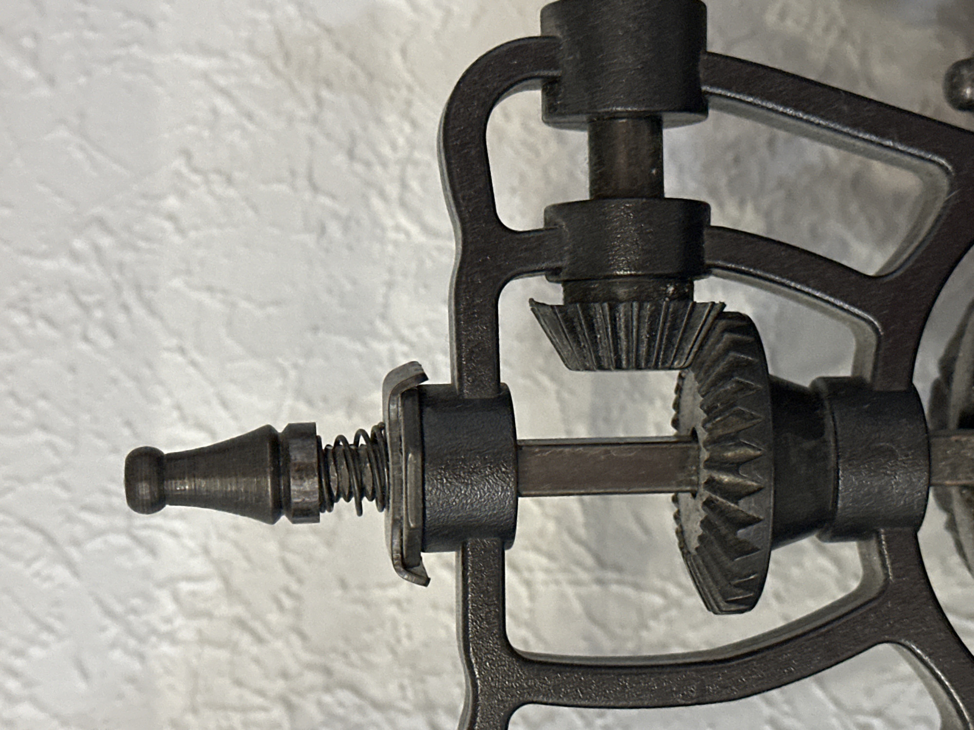 The image shows a close-up of a metal gear mechanism, likely part of a vintage hand-cranked device such as a drill or grinder. The metal is dark and appears to be cast iron or a similar material, with visible teeth on the gears. A coiled spring is also visible, positioned above a circular plate. The background is a textured white surface, possibly a wall. The image is a static, eye-level shot with no people or discernible activity. There is no information within the image to suggest a location in Nevinnomyssk, Russia.