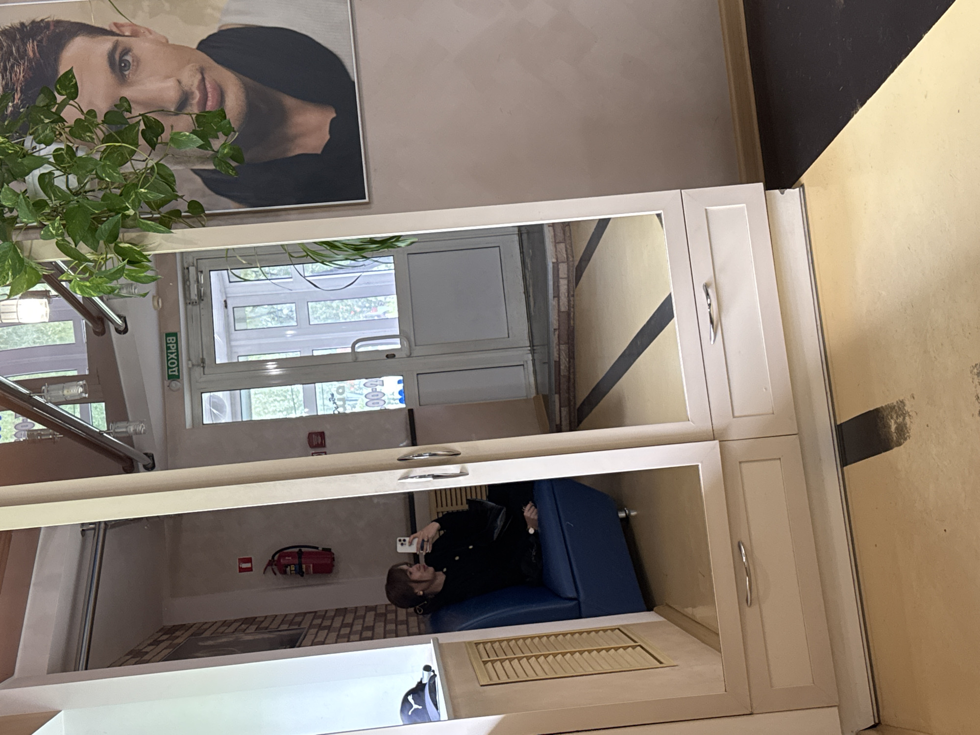 The image shows a mirror reflection of a person sitting on a blue seating area inside a building. The person is holding a smartphone, possibly taking a picture or video. A fire extinguisher is visible on the wall nearby. The location is likely in Chaikovsky, Russia, as indicated by the Cyrillic text on the exit sign visible in the mirror. The mirror is part of a cabinet or shelving unit. Above the mirror is a framed portrait and some greenery. The building has a door with multiple glass panes. The overall environment appears to be an interior space with a modern design, potentially a restaurant or public space.