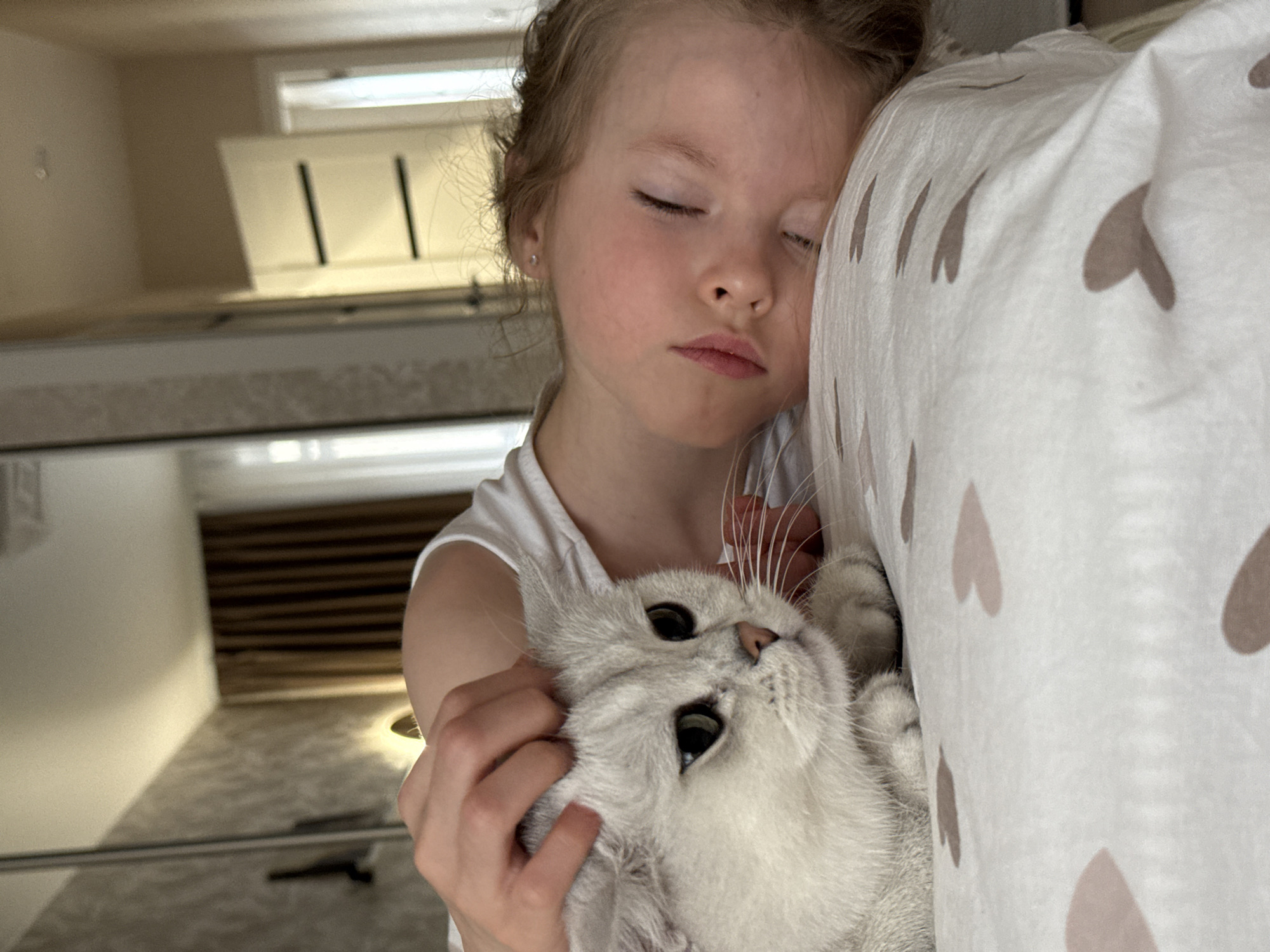 A young girl with fair skin and closed eyes is holding a white cat with large, dark eyes. The girl is wearing a white t-shirt and has light brown hair pulled back. Her face is relaxed, suggesting sleep or rest. The cat is nestled in her arms, its whiskers prominent. The background includes a white pillow with brown heart patterns, and out-of-focus shelves or storage units. The location in the city of Izhevsk, Russia is not discernible from the image.