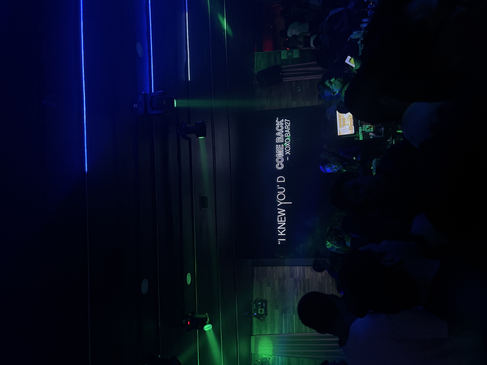 The image depicts the interior of a venue, likely a bar or club, in Puebla, Mexico. The scene is dimly lit with blue and green lighting creating a night-time atmosphere. On the left, vertical blue lines illuminate a wall, while moving head lights with green beams are pointed across the room. Towards the center of the image, a dark wall displays the text ""I KNEW YOU'D COME BACK"" - XOXO.BAR27" in a bright font. A crowd of people, mostly silhouetted in the darkness, is present. Some individuals are standing and facing the stage or display. The flooring appears to be made of wood.