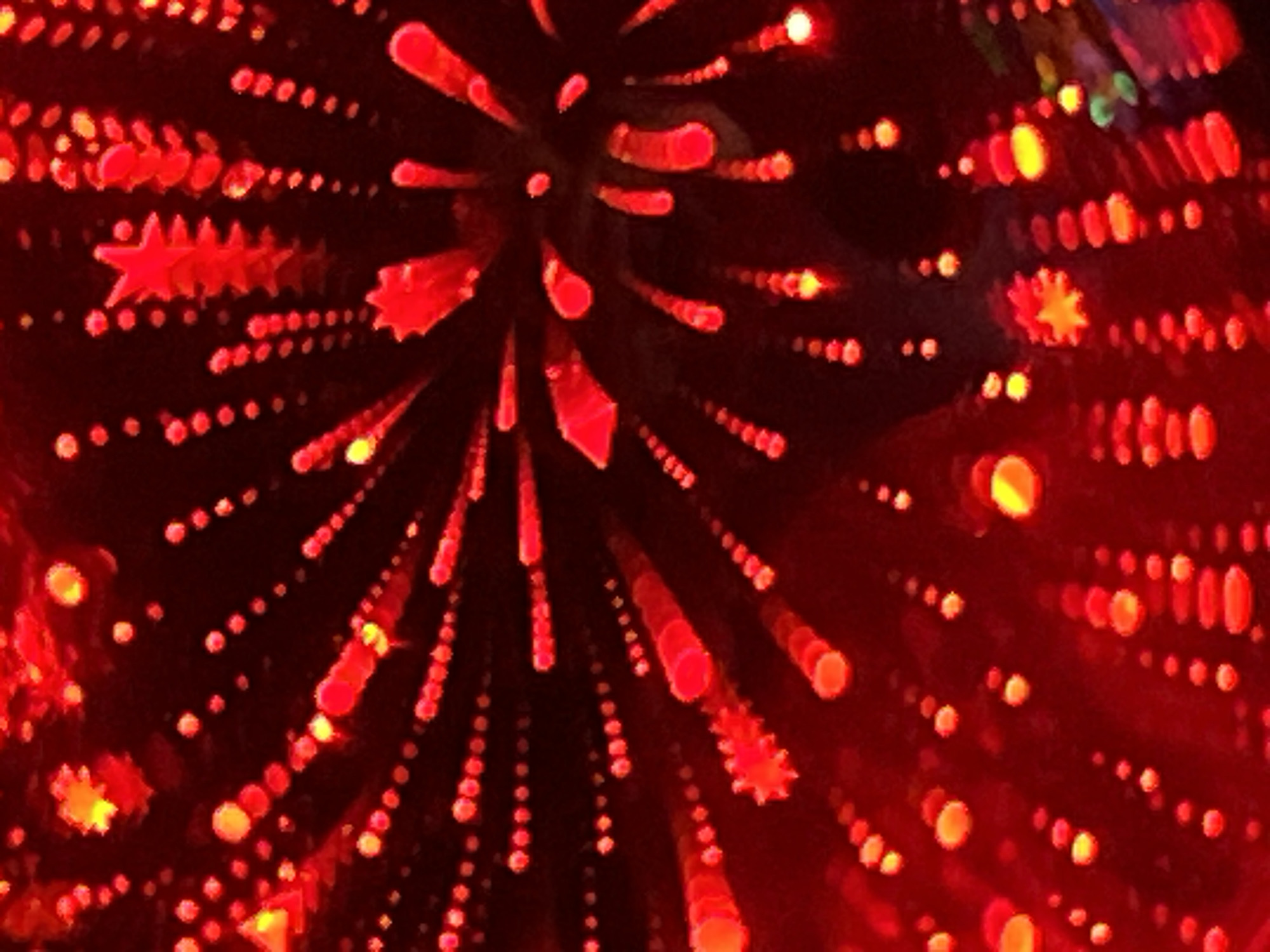 The image is a close-up, abstract shot of red and yellow lights against a dark background. The lights are out of focus, creating a bokeh effect with distinct circular and star shapes. The lights appear to be arranged in radiating patterns, resembling fireworks or light trails. Some of the lights are small and clustered together, forming lines or sprays, while others are larger and more distinct. The overall impression is one of a vibrant and energetic display of light. The scene appears to be indoors, with no visible environmental context suggesting a specific city or location.