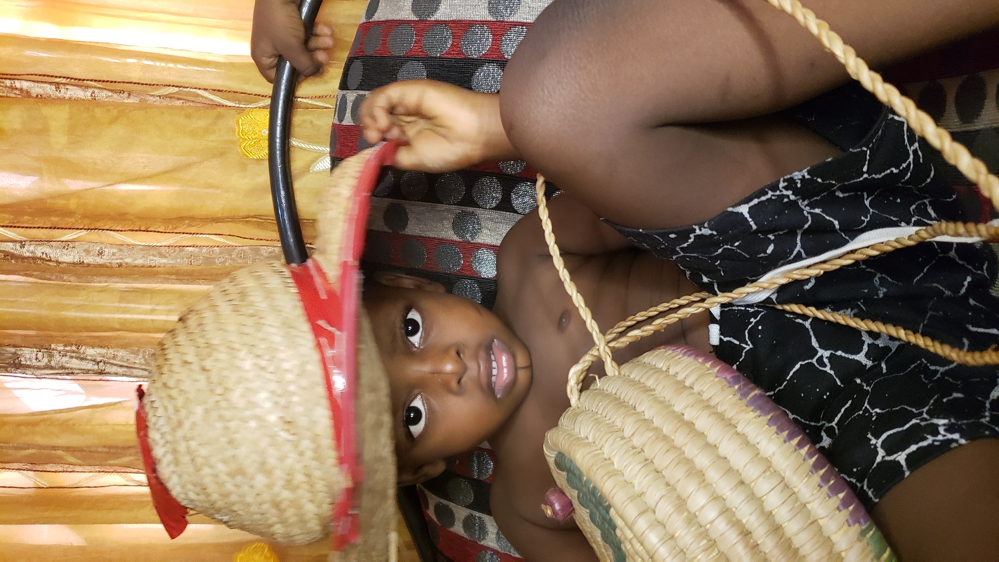 In Sabon Tasha, Nigeria, a young boy is seated on a patterned couch. He is wearing a straw hat adorned with a red ribbon, which is tilted over his forehead, partially obscuring his eyes. The boy is holding a woven basket in his lap and appears to be looking directly at the camera. His chest is bare, and he is wearing black and white patterned shorts. Behind him, a yellow, translucent curtain hangs. A black metal bar, possibly part of the couch or a nearby railing, is visible in the upper right corner of the frame, with a hand reaching for it. The lighting suggests an indoor setting, possibly during the daytime due to the natural light filtering through the curtain.