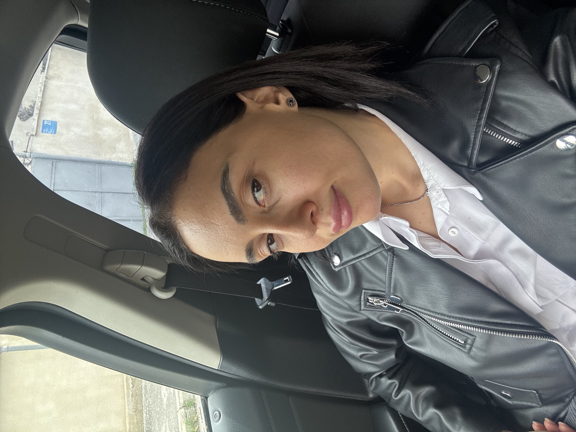 A woman is seated inside a car, wearing a black leather jacket over a white button-down shirt. The background shows a street scene through the car window, with a blue street sign visible indicating the location as Tbilisi, Georgia. The car's interior is visible, including the seat, seatbelt mechanism, and parts of the door. The woman is looking at the camera with a slight smile. She has dark hair and is wearing an earring.