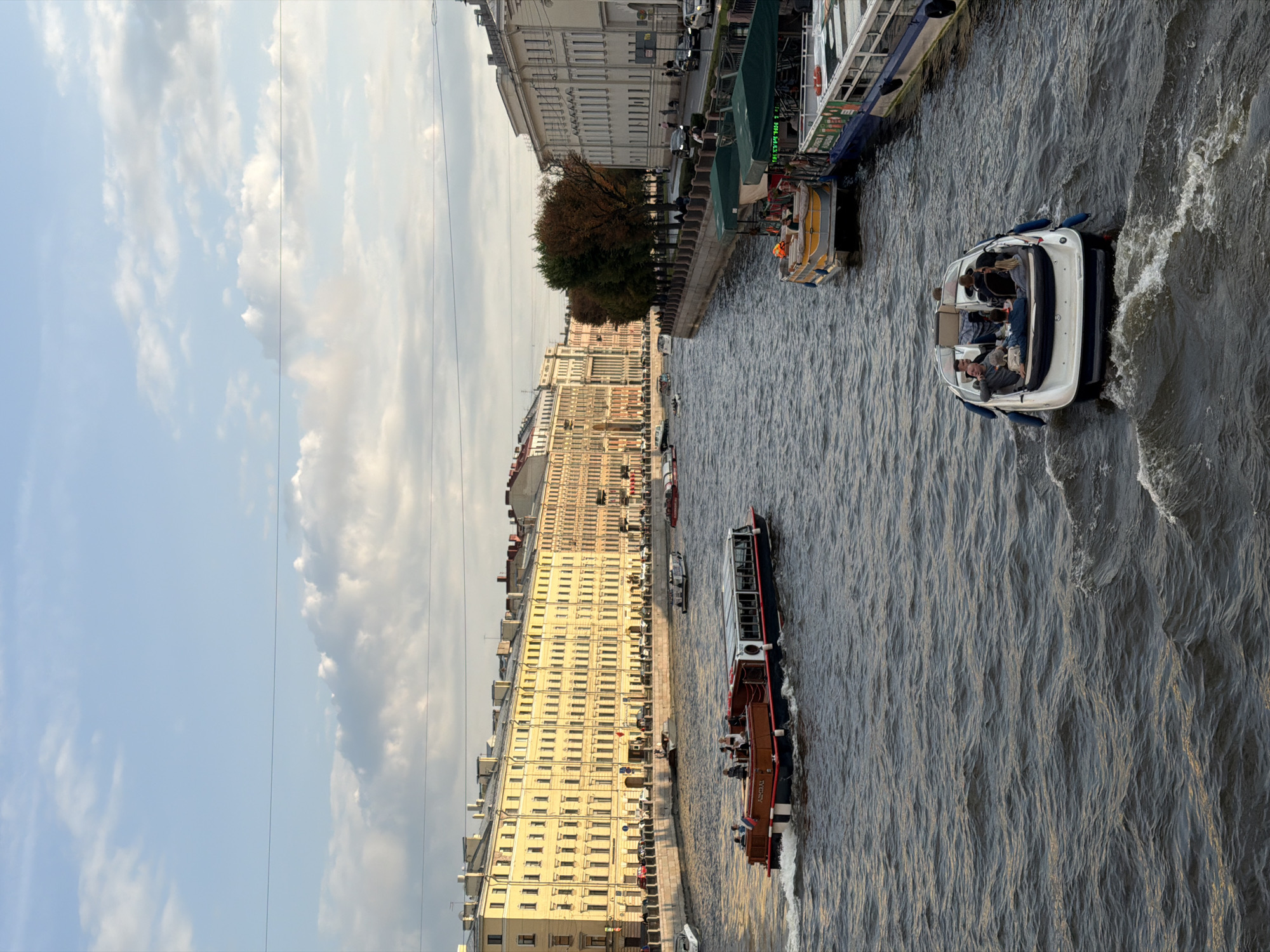The image shows a wide canal in Saint Petersburg, Russia, bordered by grand, historic buildings on both sides. Several boats are navigating the canal, some carrying passengers. In the foreground, a white motorboat with an open top carries several people who appear to be tourists enjoying the ride, with one person leaning back, relaxed. Further up the canal, a larger, more traditional tour boat named "ADMIRAL" is visible, also carrying passengers. On the left bank, a long, pastel yellow building with numerous windows and balconies lines the canal. On the right bank, a cream-colored building with similar architectural features can be seen. Trees with autumn foliage line the right bank walkway. The sky is partly cloudy with a mix of blue sky and white clouds, and the late afternoon sun casts a warm glow on the buildings. The water of the canal is choppy, with visible waves and wakes generated by the boats. The scene depicts a lively urban waterway scene with elements of tourism and historical architecture characteristic of Saint Petersburg.