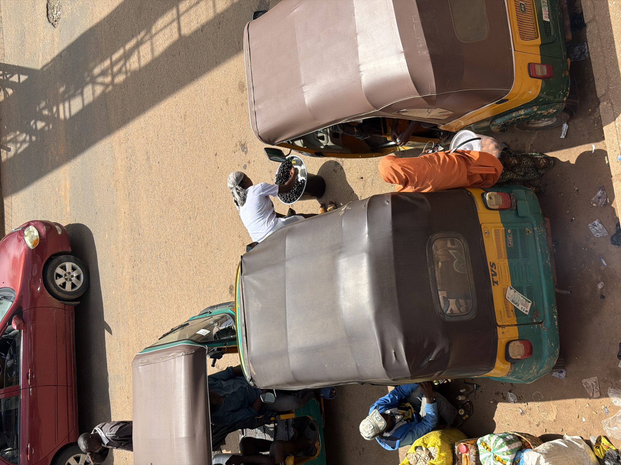 This image shows a street scene in Bukuru, Nigeria, likely during daylight hours as indicated by the strong shadows. The scene features several auto-rickshaws, commonly known as "keke" in Nigeria, a red car, and people.
In the foreground, a person in a white shirt and headscarf is holding a large metal bowl filled with dark, round objects, possibly produce or other goods. This person is standing next to a keke with a brown canopy. Another keke, with a brown and green body, is parked next to it, and a person in an orange garment is lying on top of it. Another keke with a green and yellow body is also visible.
Further down the street, a red car is parked, and a person is seen looking under it. Another person in a blue shirt is sitting on the ground near a pile of goods, including what appears to be watermelons and other produce in plastic bags. The ground is paved with asphalt and appears dusty, with some scattered litter.
The overall impression is of a busy marketplace or roadside area where goods are being sold or transported. The strong shadows suggest it is either morning or late afternoon. There is no visible text in the image that provides further context about the specific location or time.