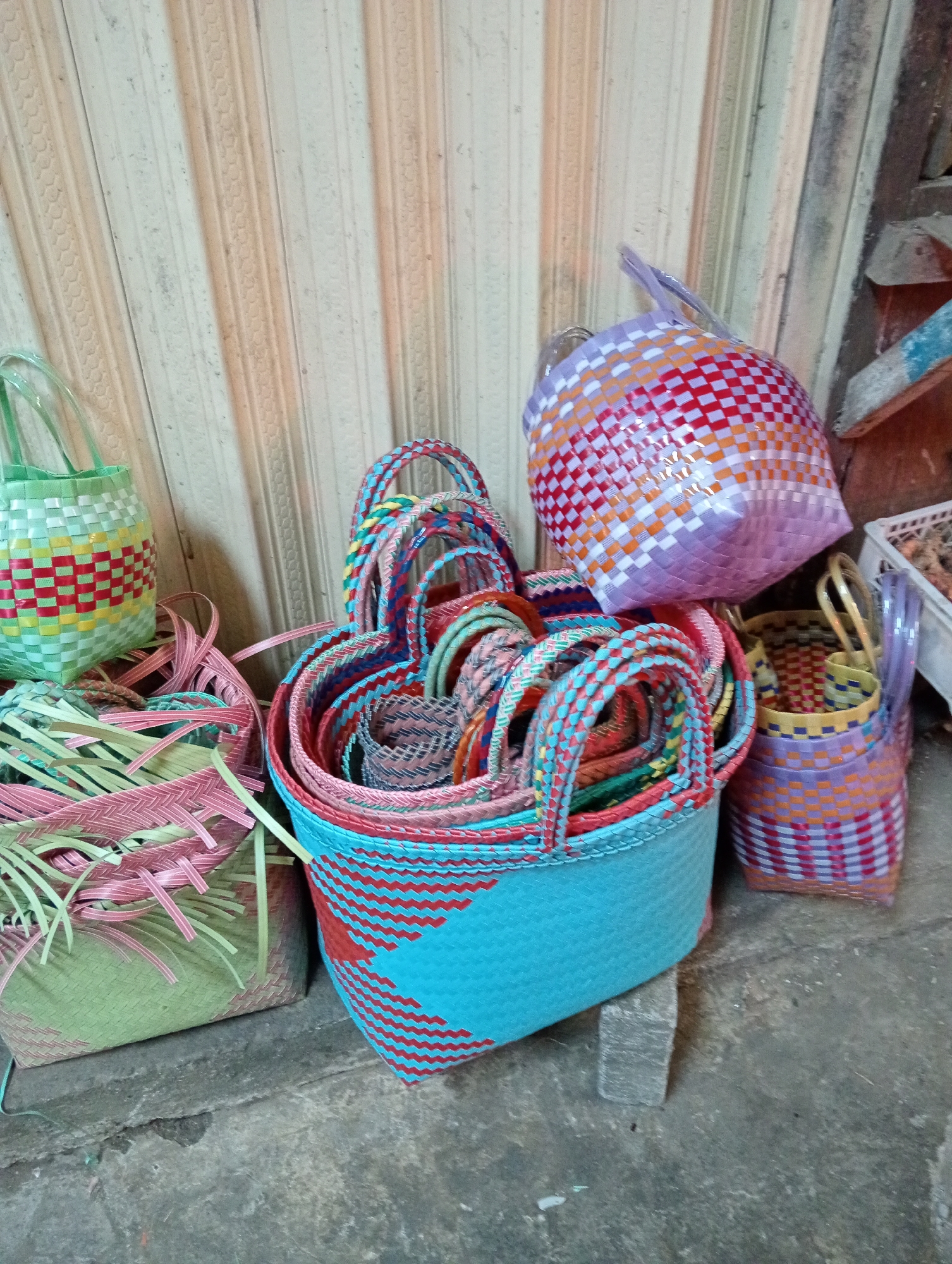 A collection of brightly colored woven plastic baskets is displayed on a concrete floor in front of a wall with vertical ribbed paneling. In the foreground, a large, teal-colored basket with red zig-zag patterns is propped up by a small grey block. To its left, a pale green basket with pink and green woven strips is overflowing with loose strips of pink and green plastic. Behind these, a light green woven basket with red and yellow checkered patterns has a clear plastic handle. To the right of the teal basket, a small, purple basket with orange and pink checkered patterns sits on the floor. Further back, a larger, round basket with purple, orange, red, and white checkered patterns is tilted forward, its handle visible at the top. The overall scene suggests a small shop or market stall, likely in Grabag, Indonesia, where these handcrafted items are for sale.
