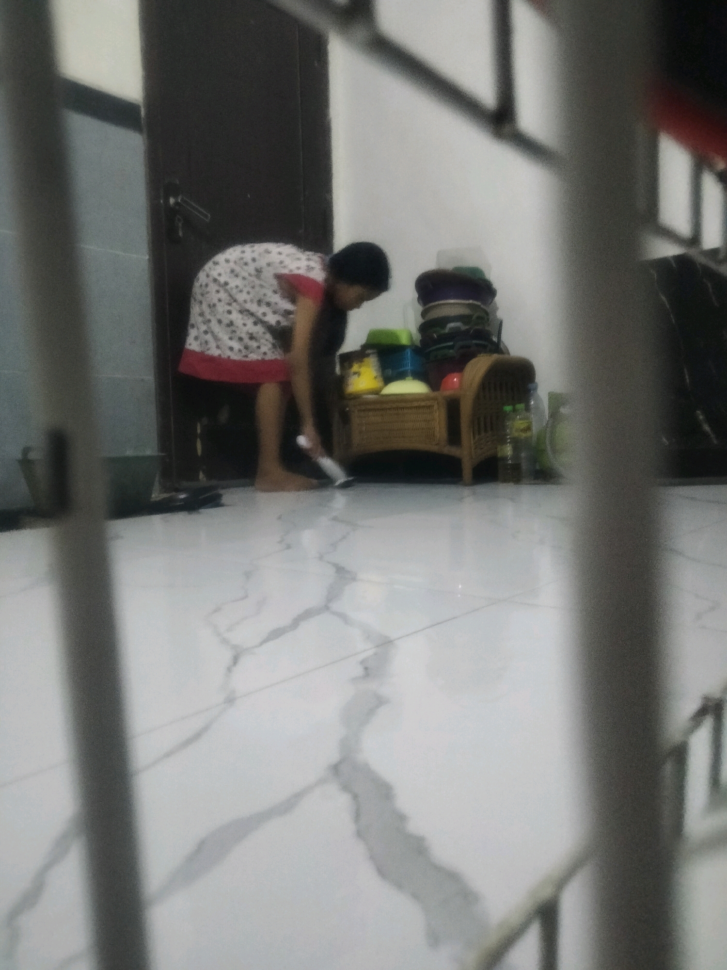 The image captures an indoor scene with a person actively engaged in cleaning. The individual, dressed in a white and red patterned garment, is stooped over, holding a white cleaning tool, likely a mop or broom, and appears to be sweeping the floor. The floor is tiled with a white marble-like pattern and shows streaks that suggest recent cleaning.
To the right of the person is a small, woven rattan furniture piece, possibly a bench or storage unit, which is holding various colorful items, including what appear to be plastic bowls or containers stacked atop one another. A few plastic bottles are also visible on the right side of the furniture.
In the background, a dark-colored door is prominent on the left, with a handle visible. Adjacent to the door is a partially obscured grey cylindrical object, possibly a bucket or bin. The walls appear to be a light color, with some darker vertical lines suggesting shadows or architectural features. The overall lighting is dim, creating a slightly hazy atmosphere. The scene takes place indoors, on a tiled floor, with elements of domestic activity. The question of the city of Soko, Indonesia, is not visually supported by the image content; therefore, no specific location can be determined from the visual information provided.