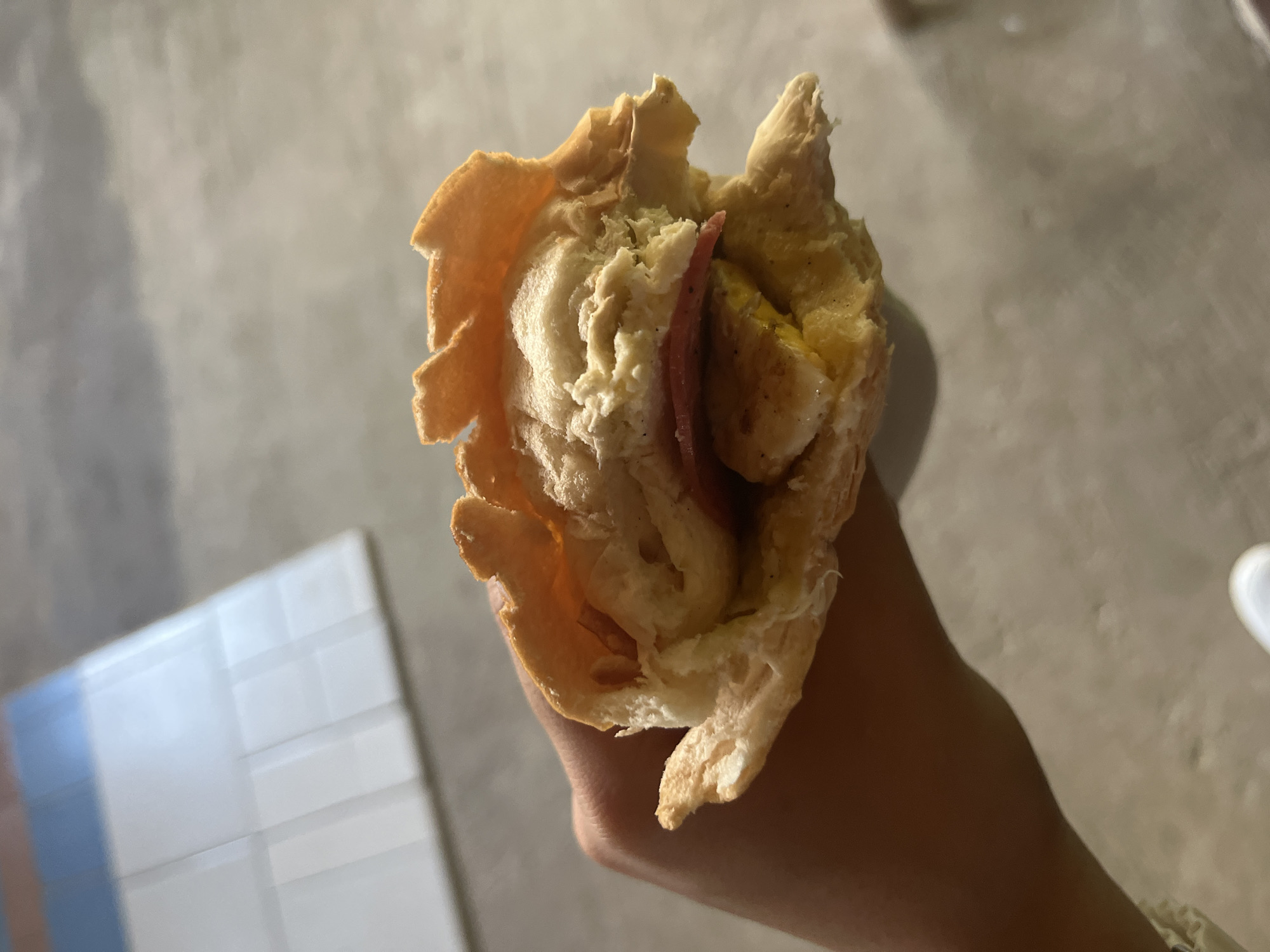 A person's hand holds a sandwich made with a puff pastry bun, filled with sliced ham and cooked egg. The background is blurred and appears to be an outdoor setting. The ground is a textured grey surface, and in the upper left corner, a tiled surface with blue and white tiles is visible. The lighting suggests it is daytime. The image was taken in Juan Griego, Venezuela.
