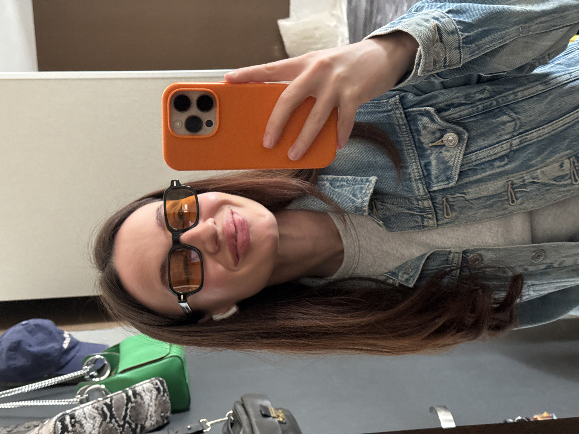 A woman is taking a selfie with an orange-cased iPhone. She is wearing a denim jacket over a grey shirt and sunglasses. An earbud is visible in her left ear. Several handbags, including a green one and a grey snake-print bag, are visible on a surface in the foreground, along with a blue baseball cap. The background appears to be an interior space with white or light-colored walls. There is no verifiable information to determine if the image was taken in Tel-Aviv, Israel.