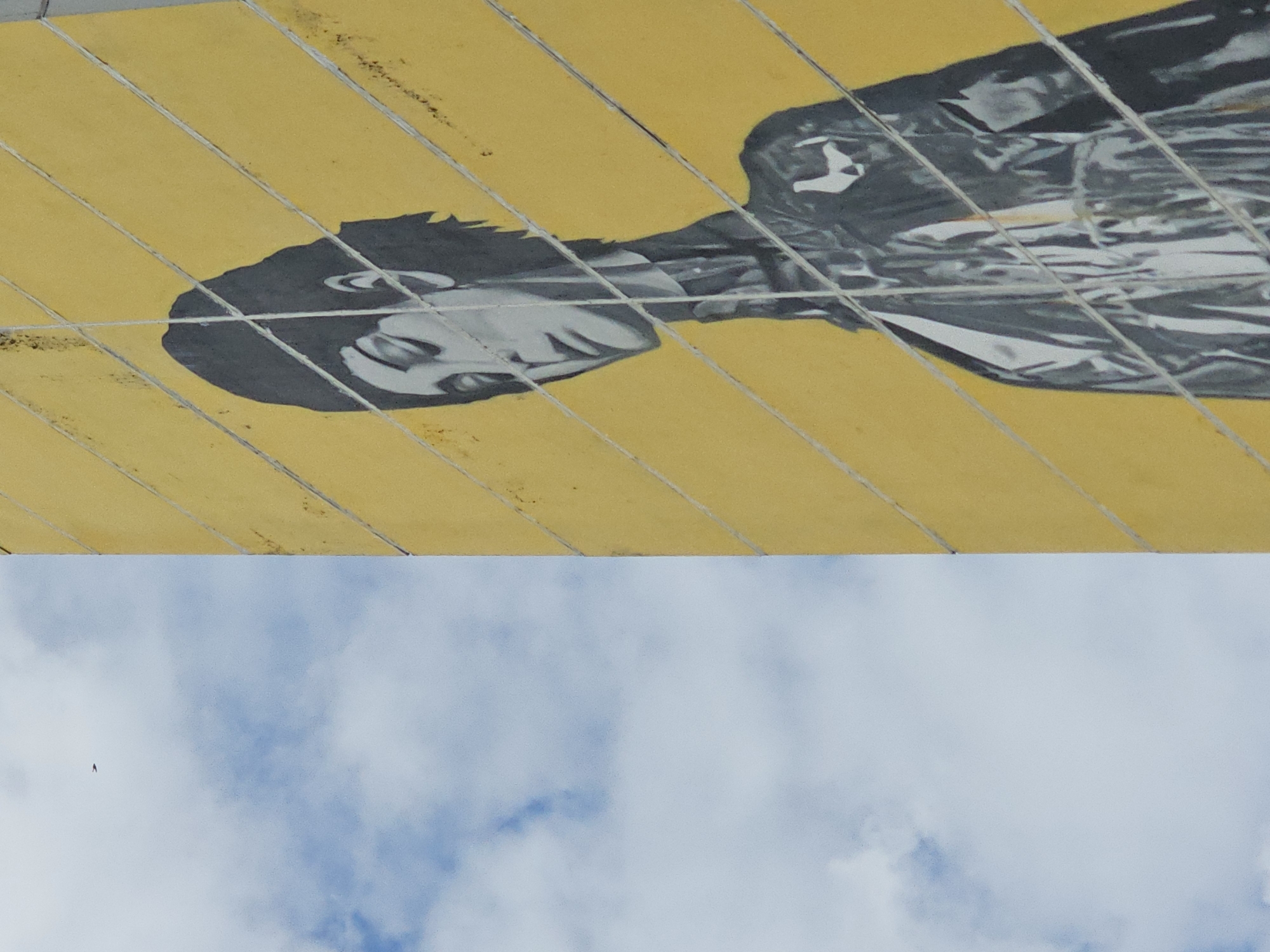 The image shows a section of a yellow wall adorned with a stencil art depiction of a person with short, dark hair, looking downwards. The artwork is rendered in shades of gray and black. The wall is divided by horizontal lines. To the left of the wall is a bright blue sky with scattered white clouds and a small dark object, possibly a bird, is visible in the sky. The composition captures the juxtaposition of the urban art against a natural backdrop. This mural is located on the facade of a residential building at 5 Molodezhnaya Street in Odintsovo, Russia.