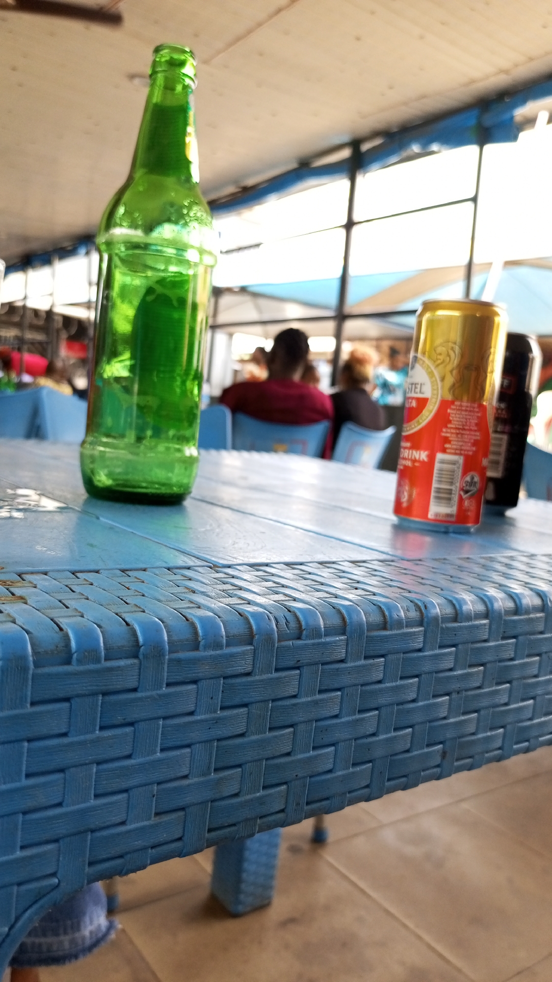 The image shows a casual outdoor scene, likely at a restaurant or bar in Sabon Tasha, Nigeria. A blue woven plastic table dominates the foreground, holding a green beer bottle, a partially visible gold and red Amstel beer can, and another dark-colored can behind it. The background features people seated at other tables, identifiable by their blurred outlines and clothing (e.g., a person in a red shirt). The environment is semi-enclosed, with what appears to be a roof or awning structure overhead. Light is bright, suggesting it's daytime.