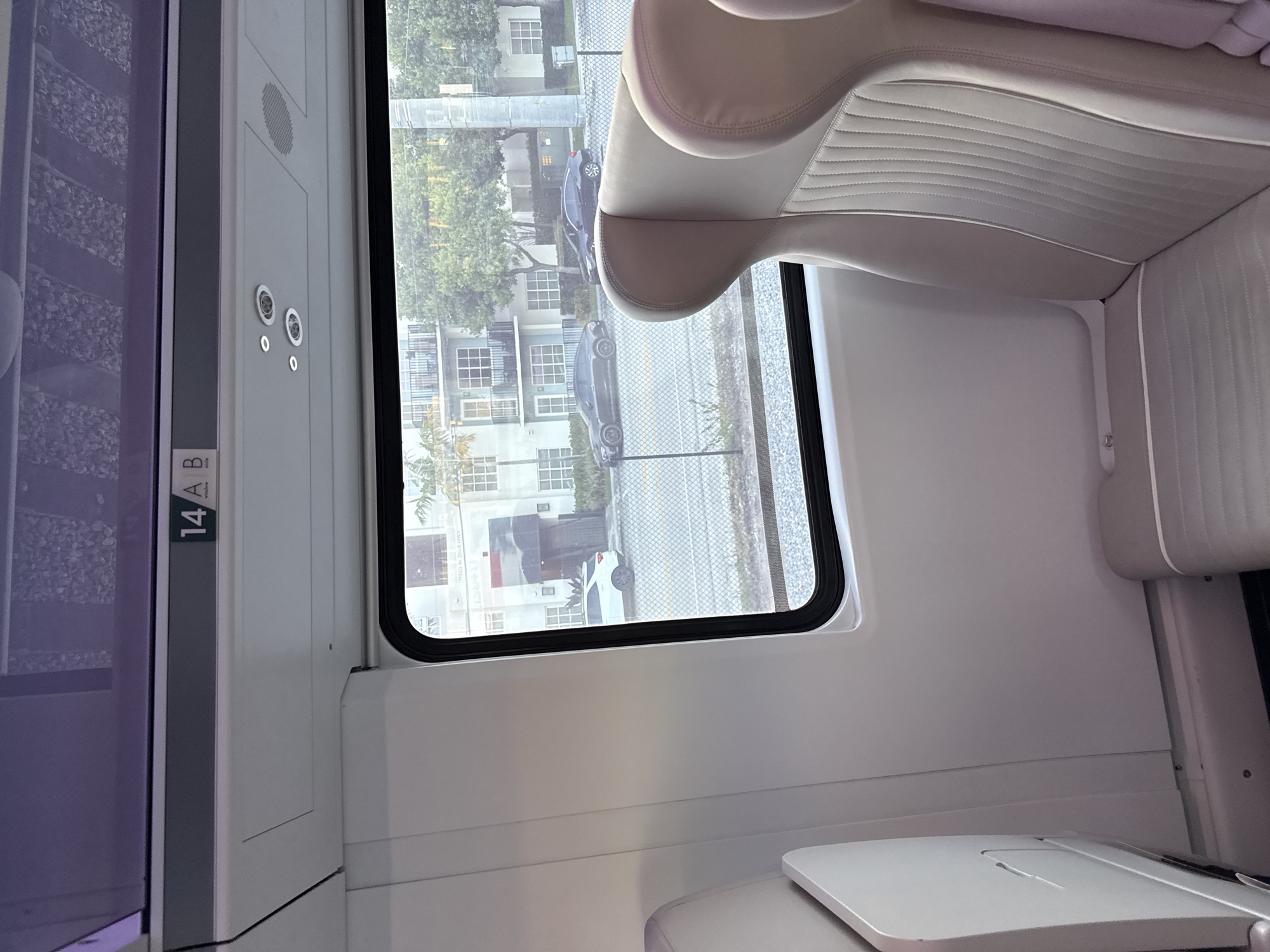 The image displays the interior of a modern train car, looking out a window at an urban setting. The train car interior features light-toned walls, a ceiling with recessed lighting and a speaker, and an overhead compartment reflecting a textured surface. Above the window, the seating area is marked "14 A B". Two unoccupied passenger seats, upholstered in light grey with vertical stitching, are partially visible on the right. A folded tray table is present on the left side of the frame.
Through the window, a chain-link fence runs parallel to the train's tracks. Beyond the fence, a paved area contains three parked vehicles: a white SUV, a dark grey sedan, and another dark sedan. In the background, a light-colored, multi-story residential building with balconies is visible, partially obscured by trees. A sign on the building or nearby structure reads "19860 W DIXIE HWY". This view is consistent with the area adjacent to the Brightline railway line near the Aventura Station, specifically looking west towards West Dixie Highway in Aventura, Florida, United States.