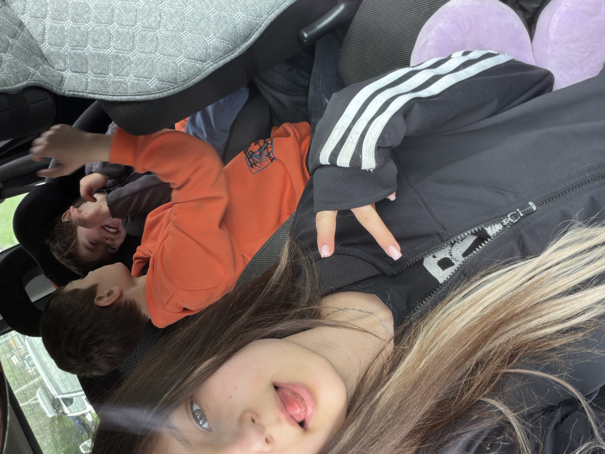 The image appears to be a selfie taken inside a vehicle, possibly a car, judging by the presence of seats and headrests. The foreground features a young woman with brown and blonde hair, making a "peace" sign with her fingers and sticking out her tongue. She is wearing a black track jacket with white stripes on the sleeves. Behind her, two young boys are visible. One is wearing an orange sweatshirt, and the other is in a grey top. The boys appear playful, with one laughing and reaching toward the camera. The surroundings are somewhat cluttered, with car seats and other objects partially visible. The location is indiscernible beyond being inside a vehicle.