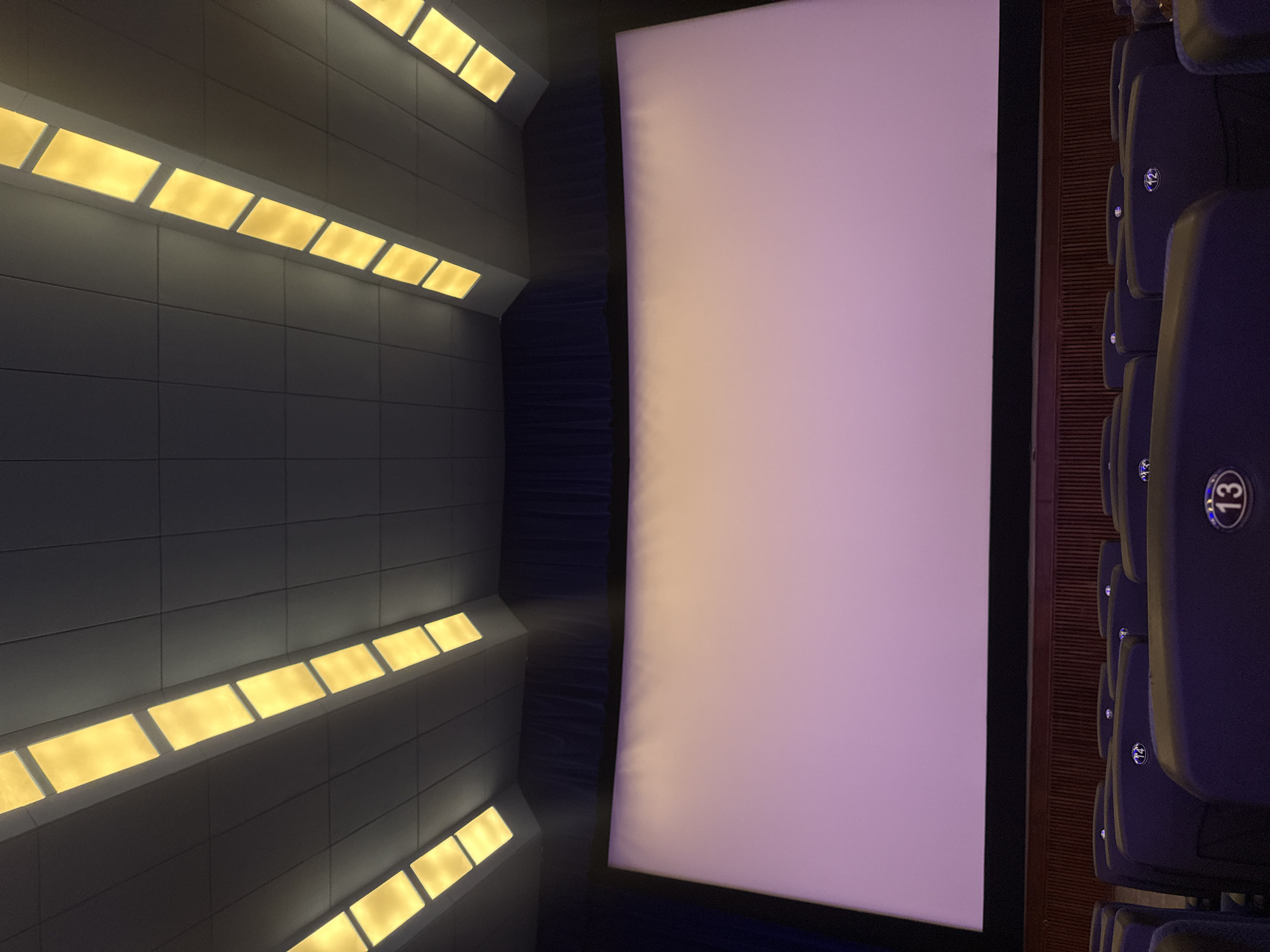 The image shows the interior of a movie theater, likely in Yerevan, Armenia. A large, rectangular white screen dominates the scene. To the right of the screen, rows of dark blue or purple upholstered theater seats are visible, with seat numbers "12," "13," and "14" evident. The walls on the left are dark gray and lined with evenly spaced, rectangular yellow lights, creating a stark contrast. A shadowy recess behind the screen indicates the walkway to access seats. The angle suggests the photo was taken from a seat in the theater.