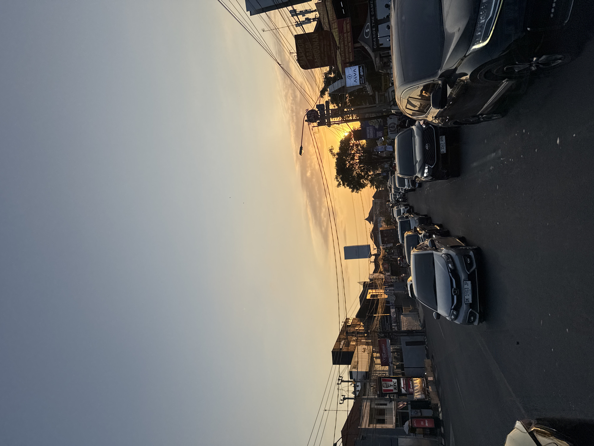 The image captures a street scene in Denpasar, Indonesia, during sunset. A traffic jam is visible on a multi-lane road lined with buildings and businesses. Several cars, including a silver Honda Brio with license plate DK 1903 JP, a black Toyota Innova with license plate B 1547 AMH, and a black SUV, are stationary or moving slowly in traffic. Overhead utility lines crisscross the sky, and streetlights are present. Businesses like KFC and others with signage in Indonesian are visible along the street. The sky is a gradient of blue at the top transitioning to orange and yellow near the horizon, indicating the setting sun which is partially obscured by trees and buildings. The overall atmosphere is that of a busy urban environment at dusk.