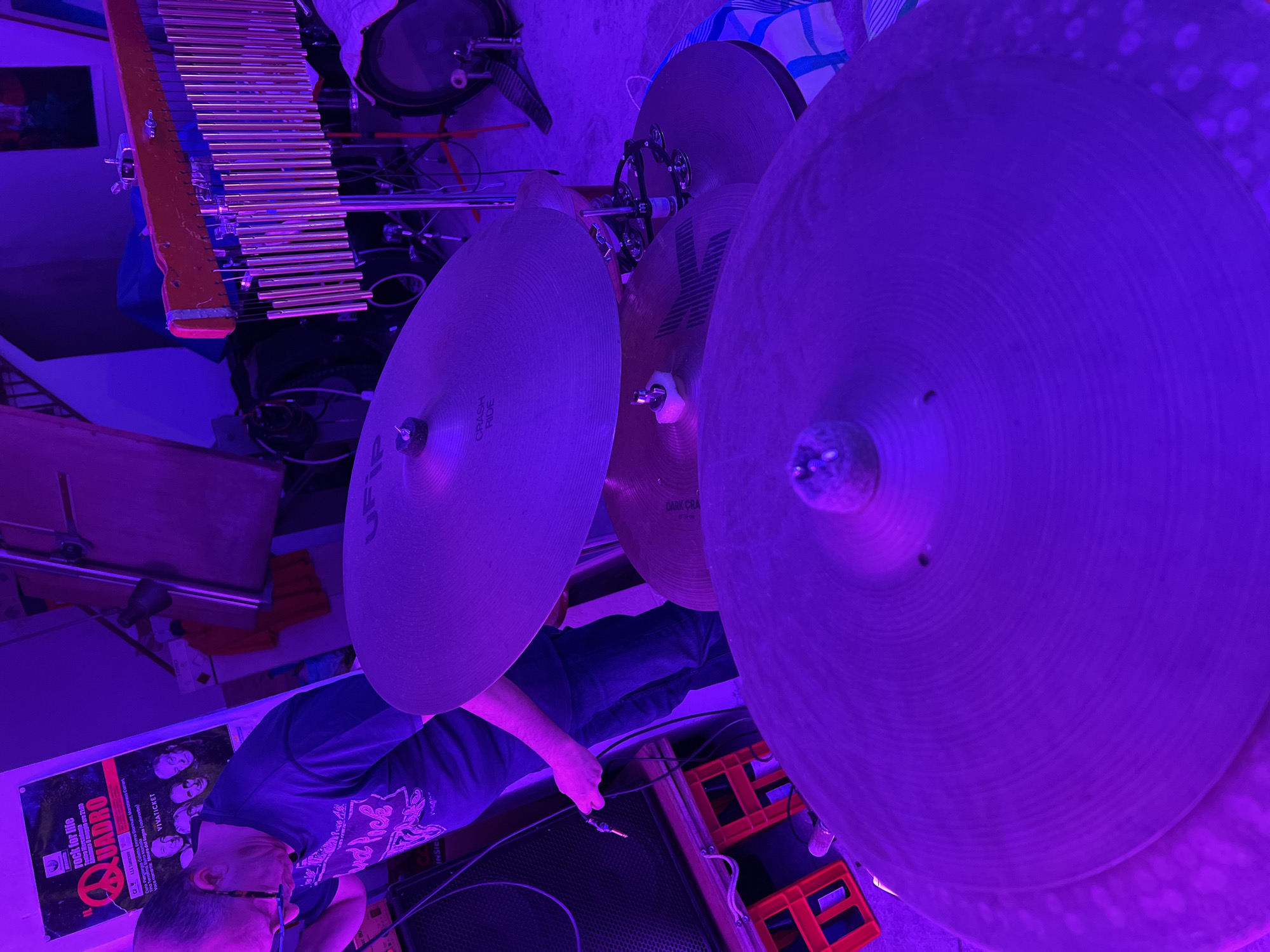 The image depicts an indoor scene in Santeramo in Colle, Italy, illuminated by a dominant purple-magenta light, characteristic of a music studio or rehearsal space. An adult Caucasian male, with short dark hair and glasses perched on his nose, is positioned on the left side of the frame. He wears a dark t-shirt with light-colored graphics and dark jeans. His head is bowed as he focuses on connecting a male 1/4 inch audio jack held in his right hand to an unseen input, likely on an amplifier or sound system, which is partially visible on the far left.
Behind the man, a poster is affixed to a light-colored wall. The poster features the text "rock for life," a peace sign, the date "Giovedì 11 maggio ore 21:00" (Thursday, May 11th, 9:00 PM), and the band name "QUADRO" in red lettering, along with musician names and "VIVATICKET."
In the midground, a drum set is partially visible from a perspective behind the cymbals. A "UFIP" brand cymbal, labeled "CRASH RIDE," is prominent, with another darker cymbal below it, partially reading "DARK CRA..." (likely Dark Crash). A small tambourine or jingle ring is mounted on a cymbal stand. To the right of the man, a set of silver-colored bar chimes, suspended from a red wooden bar, stands vertically. In the background, parts of a bass drum with a pedal are visible on the right.
The environment includes orange plastic crates stacked below the speaker on the left and a drum stool covered with a blue and white striped fabric on the right. Various cables and wires are visible connecting the equipment. The floor appears to be a hard, industrial surface.
