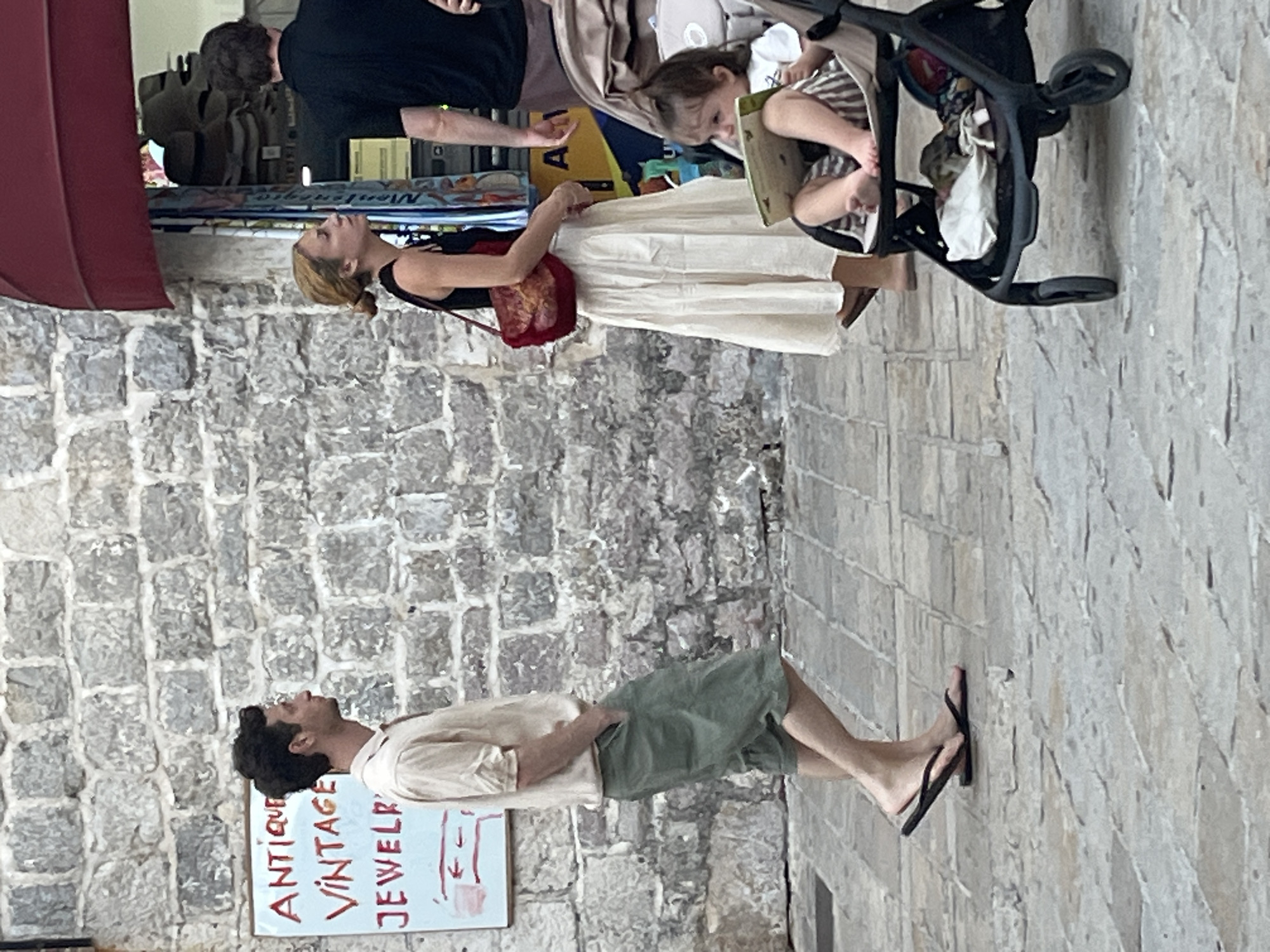 A man walks past a stone wall with a sign that reads "Antique Vintage Jewelry." He wears a light-colored, short-sleeved shirt and green shorts, with black flip-flops. To his right, a woman in a black tank top and a long, cream-colored skirt stands with a red purse. Beside her, a baby sits in a stroller, looking at a book. Behind them, a man in a black shirt stands near a storefront with hats displayed. The scene is set on a cobblestone street in Budva, Montenegro.