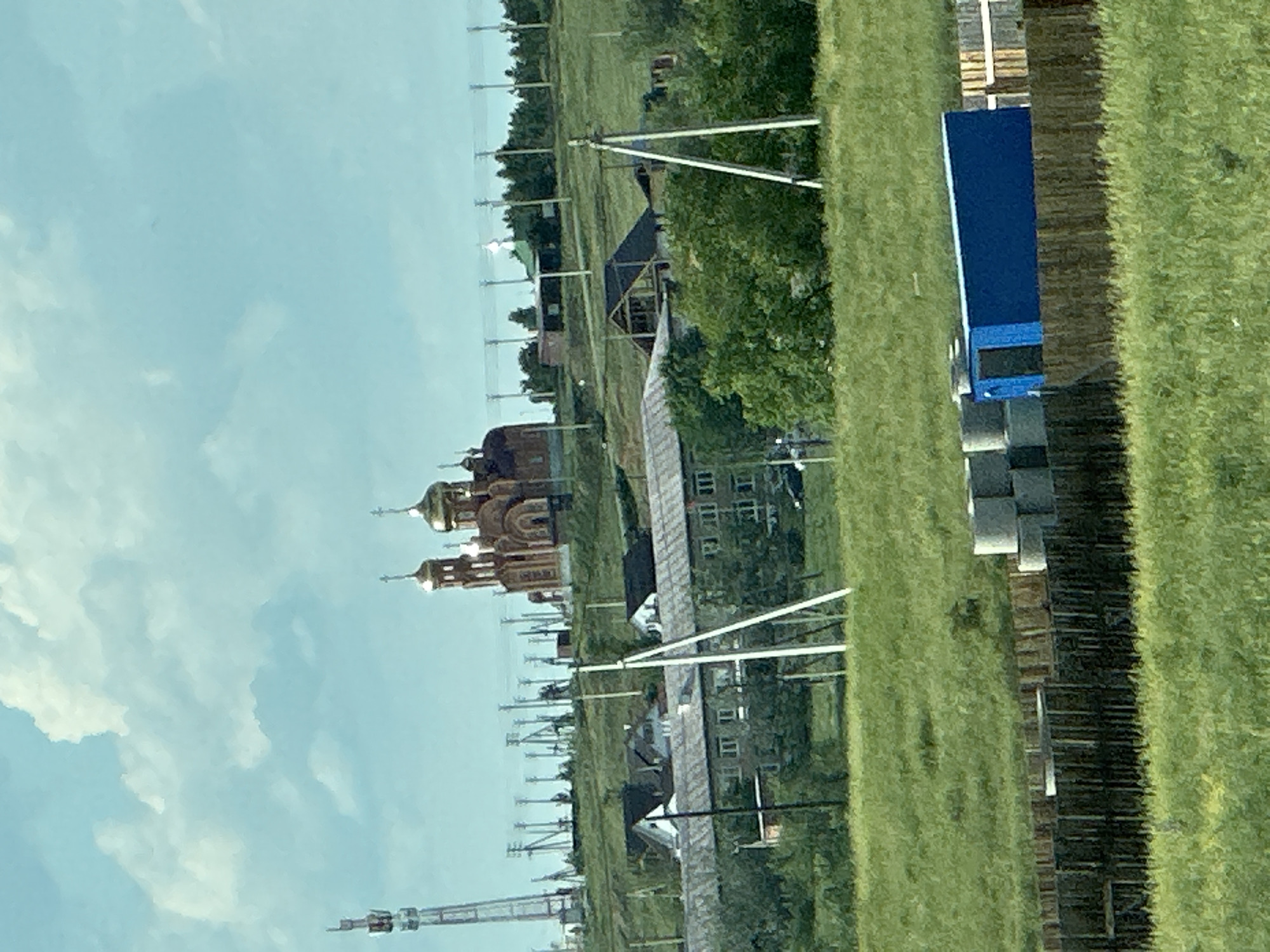 The image depicts a rural or semi-rural landscape in Борец, Russia, under a partially cloudy sky. In the background, on a hill, stands a prominent brick church with multiple golden domes. To the left of the church, a tall, slender radio tower is visible. A collection of smaller buildings, appearing to be residential or utilitarian, are scattered across the middle ground, with some nestled amongst trees. The foreground is dominated by a grassy field with a dark wooden fence running horizontally across it. In front of the fence, several large concrete rings are stacked, and to their right, a bright blue shipping container is placed. Several utility poles with crossbars are scattered throughout the scene, suggesting infrastructure for power or communication. The overall impression is of a somewhat undeveloped area with a mix of religious, residential, and industrial elements.