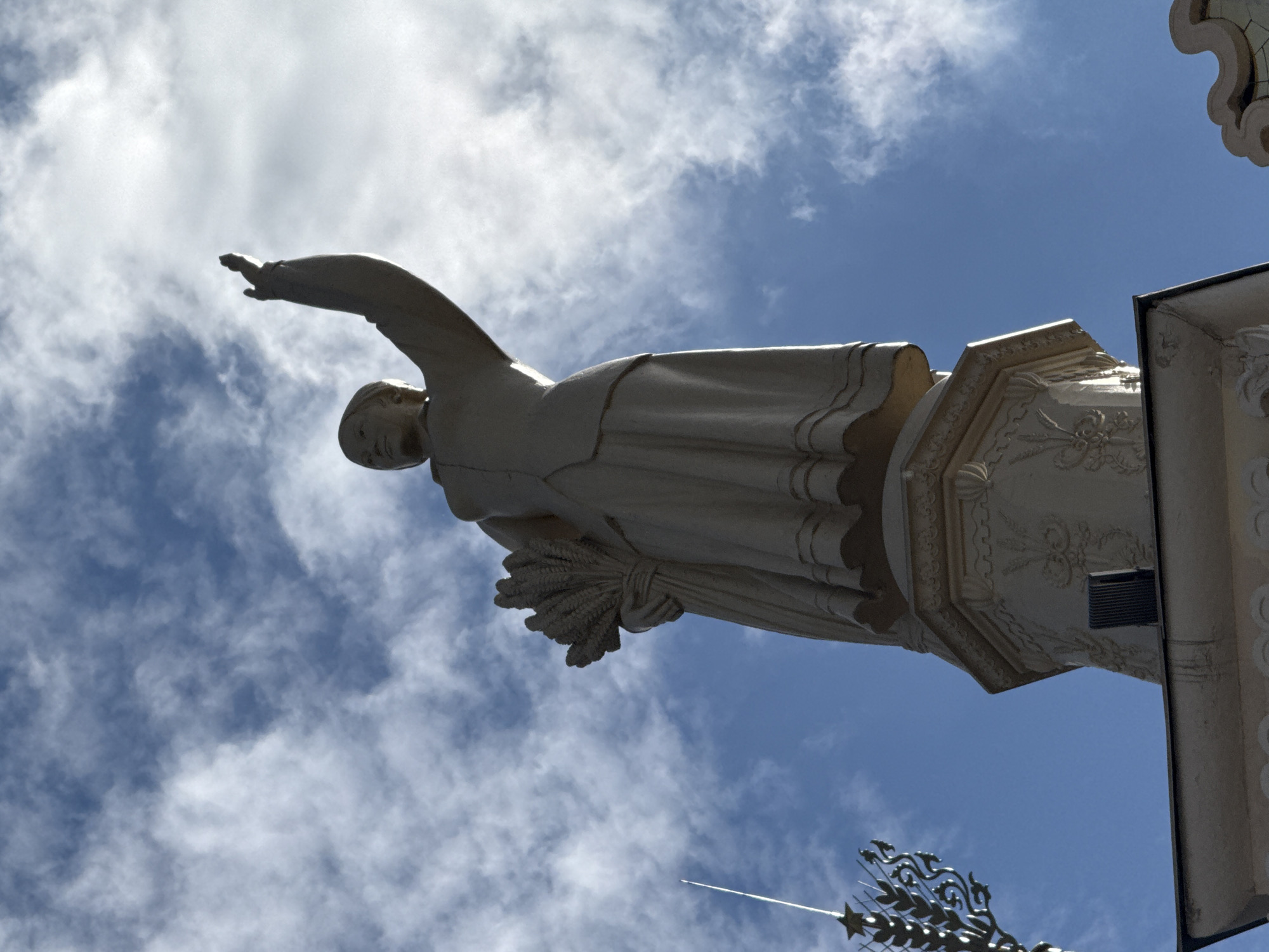 The image shows a low-angle shot of a statue of a woman against a bright blue sky filled with scattered white clouds. The statue appears to be made of a light-colored material, possibly stone or concrete, and depicts the woman with her right arm extended outwards and slightly upwards, and her left arm holding a sheaf of wheat. She is wearing a long, flowing robe that is draped in folds. The statue is mounted on an ornate pedestal with decorative carvings. The lower part of the image shows a glimpse of another architectural element, possibly part of the same structure, with detailed carvings and a dark rectangular vent. In the bottom right corner, a decorative metal element resembling a star or crest is visible.
The scene suggests a daytime setting, likely a sunny day with some cloud cover, given the bright lighting and blue sky. The low-angle perspective emphasizes the grandeur of the statue and its placement against the sky. The statue's pose and the sheaf of wheat suggest a theme related to harvest, abundance, or agriculture. Without further context or specific identifying features, the exact location of the statue is difficult to pinpoint, but the mention of Moscow, Russia, as a location context could imply a monument or artwork within the city. There is no visible text in the image.
