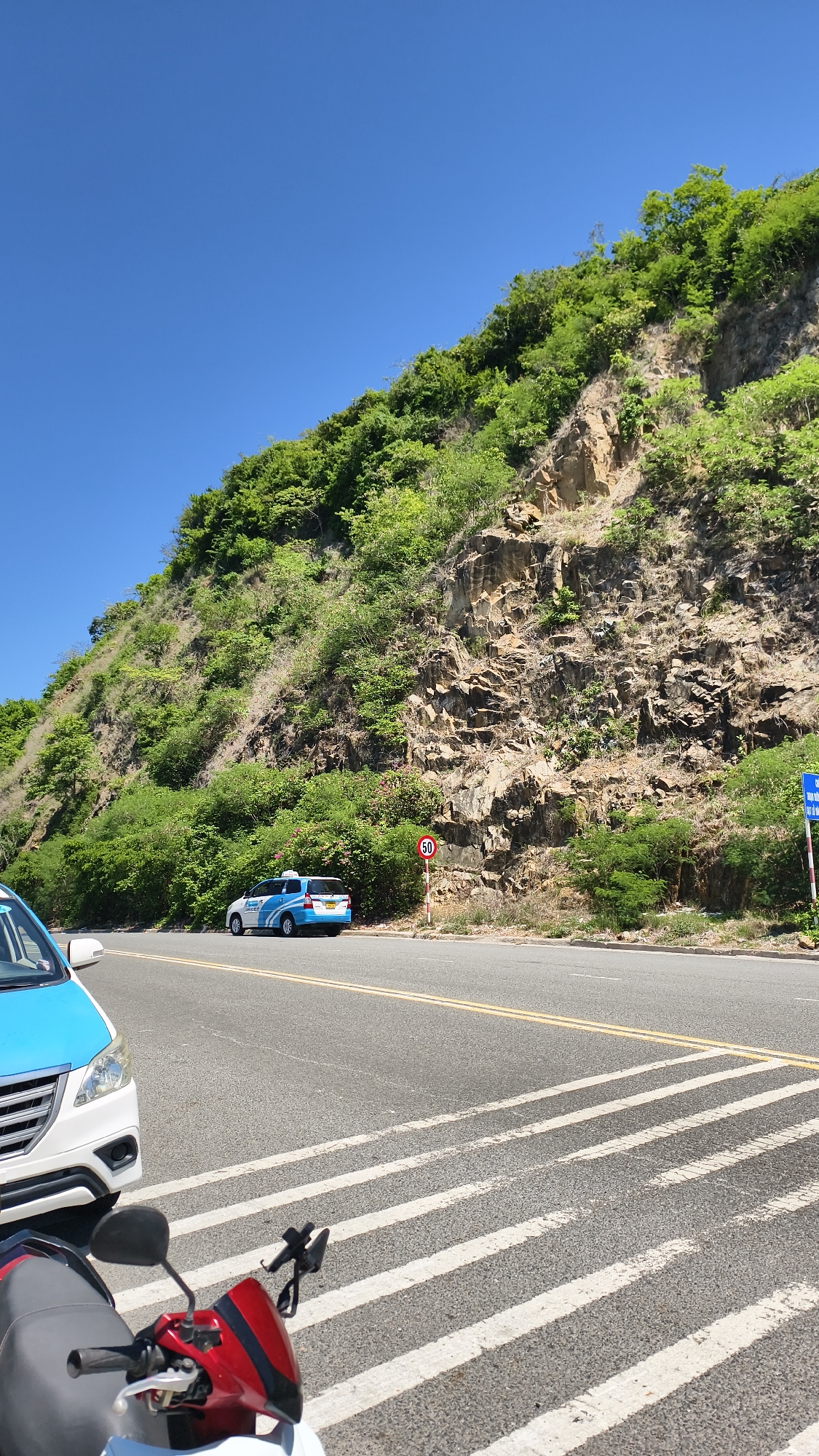 The image depicts a roadside scene on a sunny day, likely in Nha Trang Ward, Vietnam, characterized by a steep, rocky mountainside covered in lush green vegetation. A paved road runs horizontally across the lower portion of the frame, marked with yellow dividing lines and a pedestrian crossing with white stripes. On the road, a blue and white taxi is visible, and partially in the foreground on the left, a white and blue car is parked. Further in the foreground, a red and black motorcycle is parked, facing away from the camera. A speed limit sign indicating "50" is placed on the right side of the road. The sky above is clear and blue.
