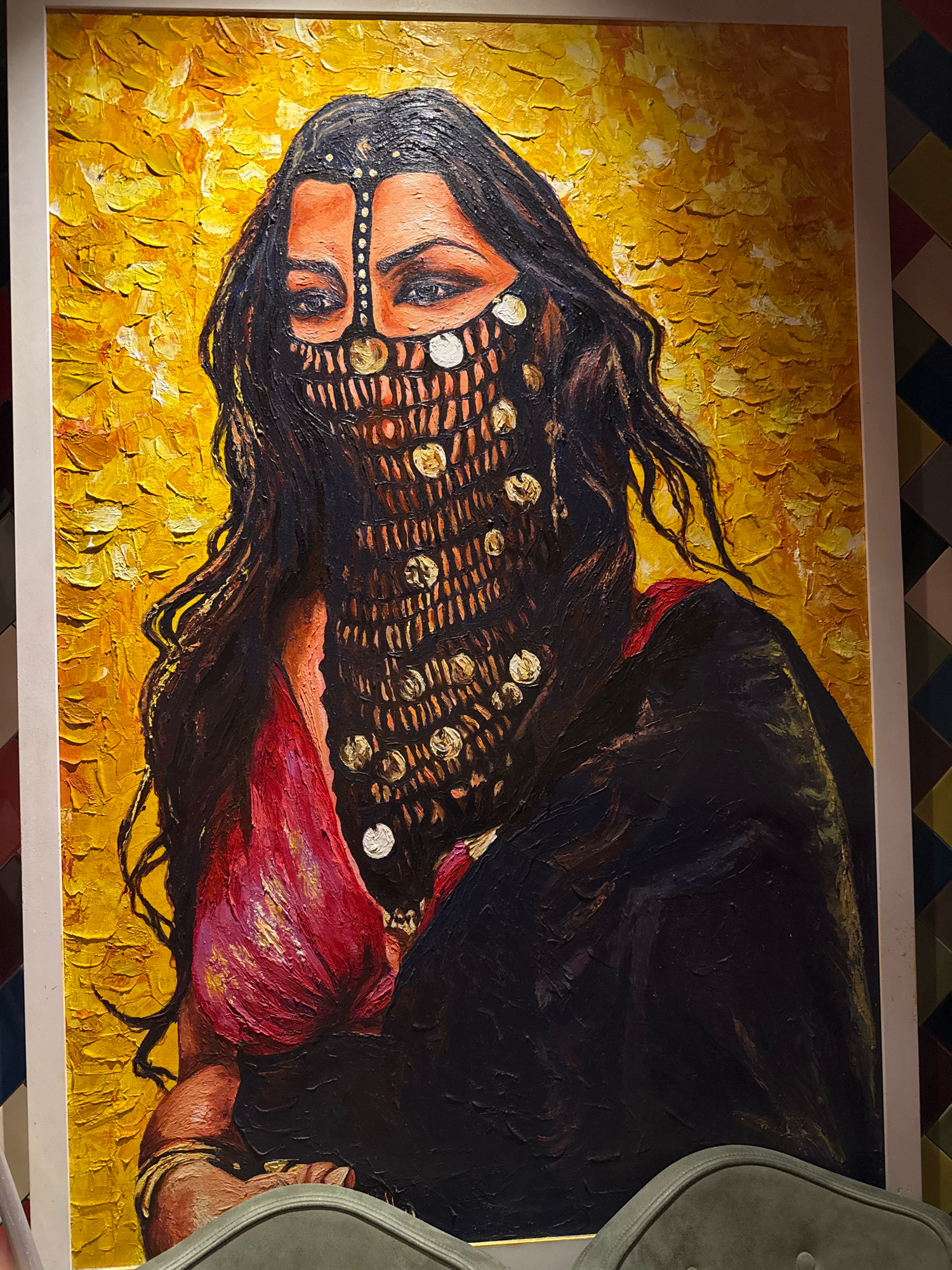 The image is a close-up, vertically oriented, full-body portrait painting of a woman with dark hair, wearing a traditional veiled headdress adorned with gold coins, a red garment, and a black robe. She is depicted against a textured, bright yellow background. The painting appears to be oil on canvas, with thick impasto brushstrokes creating a rich texture. The woman's eyes are visible through the veil, and her expression is direct and intense. The composition is centered on the woman, with the background providing a vibrant contrast to her dark attire. The painting is framed and displayed on a wall with a patterned wallpaper in the background. The location is specified as Dubai, United Arab Emirates.