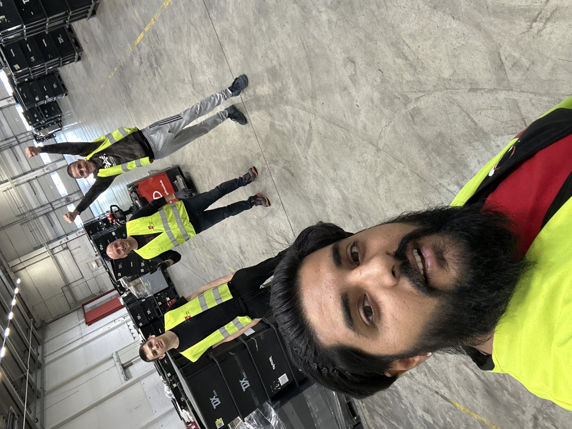 In a warehouse environment in Schweitenkirchen, Germany, three men are visible. In the foreground, a man with dark hair and a beard wearing a bright yellow safety vest over a red shirt is taking a selfie. Behind him, two other men, also wearing yellow safety vests, are in the background. One is looking at the camera, while the other has his arms raised in a celebratory gesture. Stacks of black storage containers with "TDX EUROPE" branding are visible in the background, suggesting a logistics or distribution facility. A red pallet jack is also present. The floor is concrete, and overhead lighting illuminates the space.