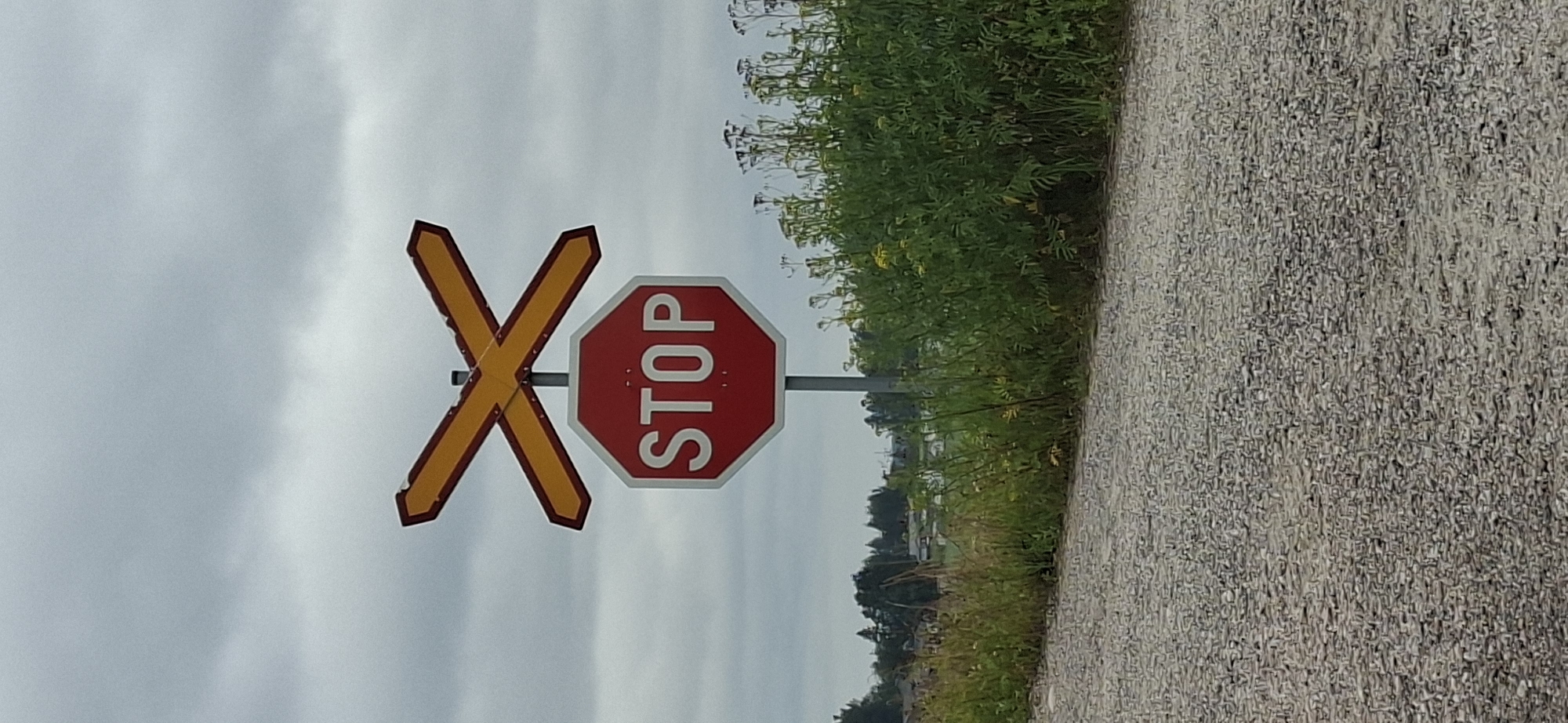 The image depicts a road sign indicating a railway crossing in Orimattila, Finland. The signpost features two yellow 'X' shaped signs stacked above a red octagonal STOP sign. The STOP sign has white text and two small white dots above and below the text. The road surface in the foreground is gravel. Behind the sign, there is a line of green vegetation and trees in the background, under a cloudy sky. There are no people or vehicles visible in the frame.