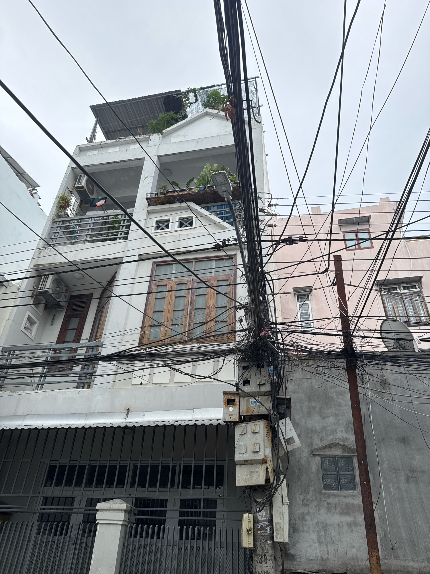The image depicts a dense tangle of electrical wires and utility poles in front of two apartment buildings. The building on the left is white and appears to be several stories tall, with balconies and air conditioning units visible. The building on the right is a pale pink color and also has multiple stories with windows and a satellite dish. The sky is overcast and the overall scene suggests an urban environment. The presence of numerous exposed wires and complex circuitry indicates a utilitarian, possibly somewhat haphazard, approach to infrastructure in this area. There are no people visible in the image. The architecture of the buildings suggests a Southeast Asian setting, consistent with Nha Trang, Vietnam, known for its densely populated urban areas with exposed electrical infrastructure. The image captures a common sight in many developing cities where utility lines are often strung together in a complex and sometimes chaotic manner.