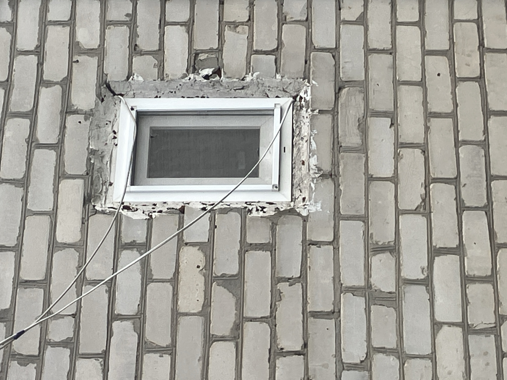 The image depicts a light-colored brick wall with a small, open window. The window frame is white and appears to be made of plastic. The area around the window is rough and appears to be under construction or in disrepair, with exposed mortar and some crumbling brickwork. Two thin, silver wires are visible, running diagonally across the wall and passing over the window. The wall itself is constructed from rectangular bricks laid in a stretcher bond pattern. The bricks are light in color, possibly white or a very pale gray, and show some signs of weathering or age, with minor chips and discoloration. The mortar between the bricks is a darker gray. The overall environment suggests an outdoor urban setting in Vovkivka, Ukraine, potentially a residential or commercial building undergoing renovation or exhibiting signs of wear. There are no people or animals present in the image.