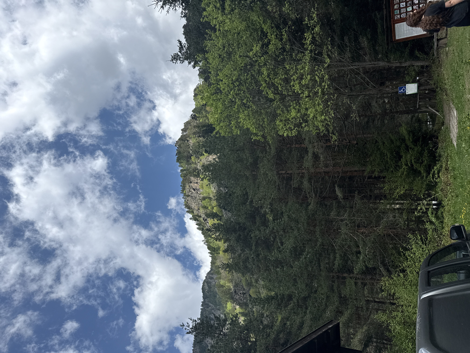 The image depicts a mountainous landscape near Beli Iskar, Bulgaria, under a sky with scattered clouds. A forested slope rises towards a rocky peak. A sign board and a blue handicap sign are present on the right edge of the image near a person. Part of a vehicle is visible in the foreground.