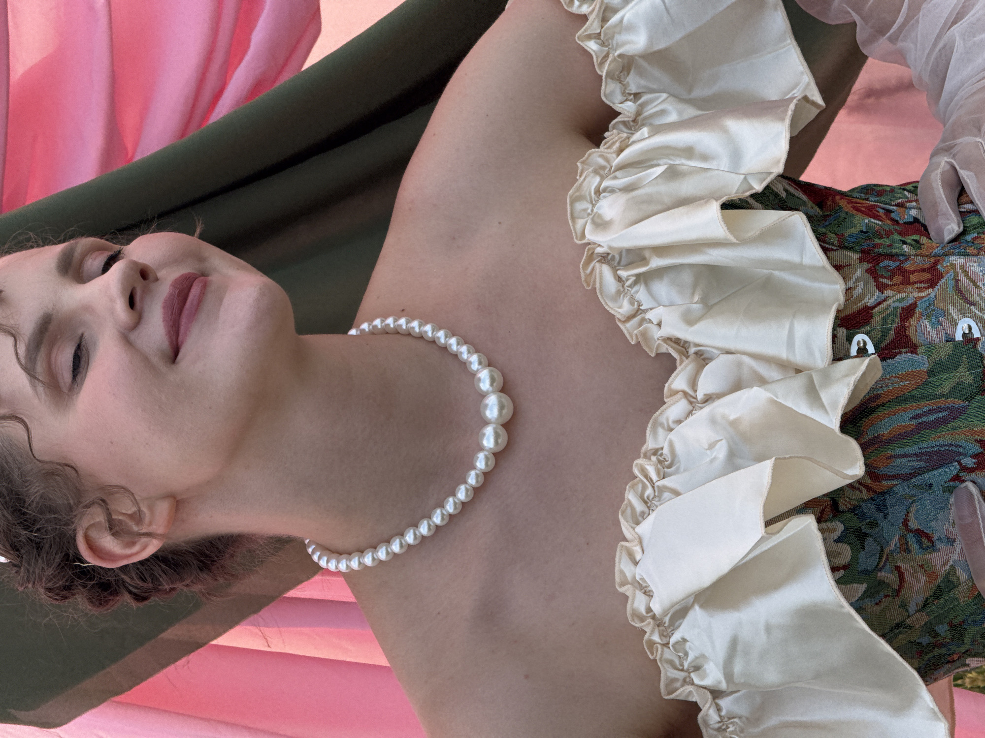 The image shows a close-up, chest-up view of a person wearing a pearl necklace and an off-the-shoulder garment with ruffled fabric. The person's hair is styled in loose curls. The background consists of draped fabric in shades of pink and green. The location is identified as Foki, Russia.