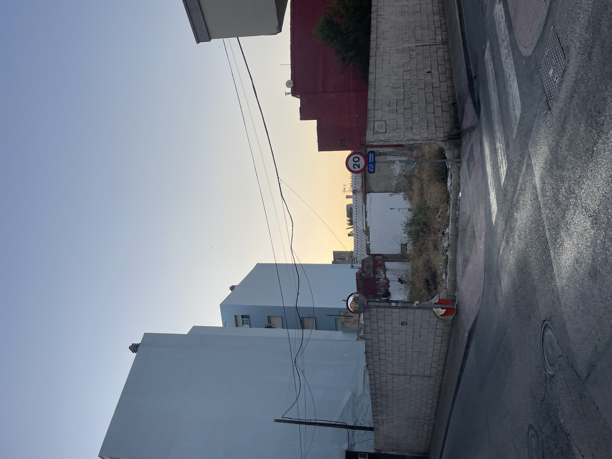 In La Línea De La Concepción, Spain, at dusk, a view of an intersection is captured. A low, concrete block wall runs along the side of the road, separating it from a vacant lot filled with dry grass and debris. Beyond the vacant lot, there are several buildings. On the left, a tall, pale blue apartment building dominates the scene, followed by a shorter, light blue building. To the right of the vacant lot is a bright red building. A speed limit sign for 20 km/h and a pedestrian crossing sign are visible near the red building. A convex mirror is mounted on a pole near the intersection, reflecting the street and buildings. The road surface is asphalt, showing signs of wear and some light reflections from the setting sun. A pedestrian crossing is marked with white stripes. The sky is a gradient of pale blue and orange, indicating either sunrise or sunset.