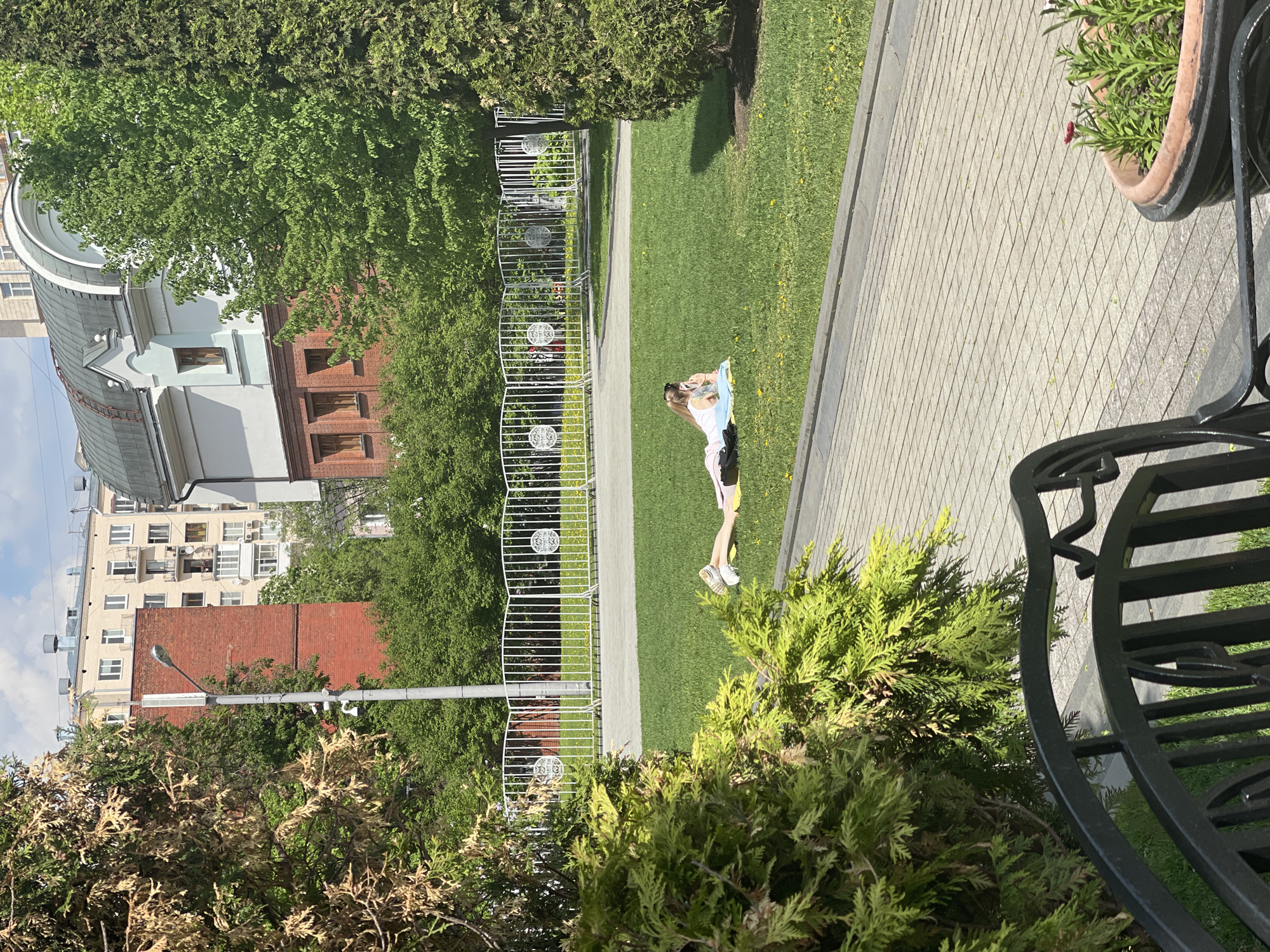 The image shows an outdoor scene, with a girl lying on a blanket on the grass in a park. The park has a white fence and a paved walkway. The area is landscaped with trees and bushes. In the background, there are buildings, including some with red brick and white facades. The scene appears to be taken from a high vantage point, possibly from a balcony or upper-story window, as a part of a black metal bench and a potted plant are visible in the foreground. The weather seems sunny and bright.