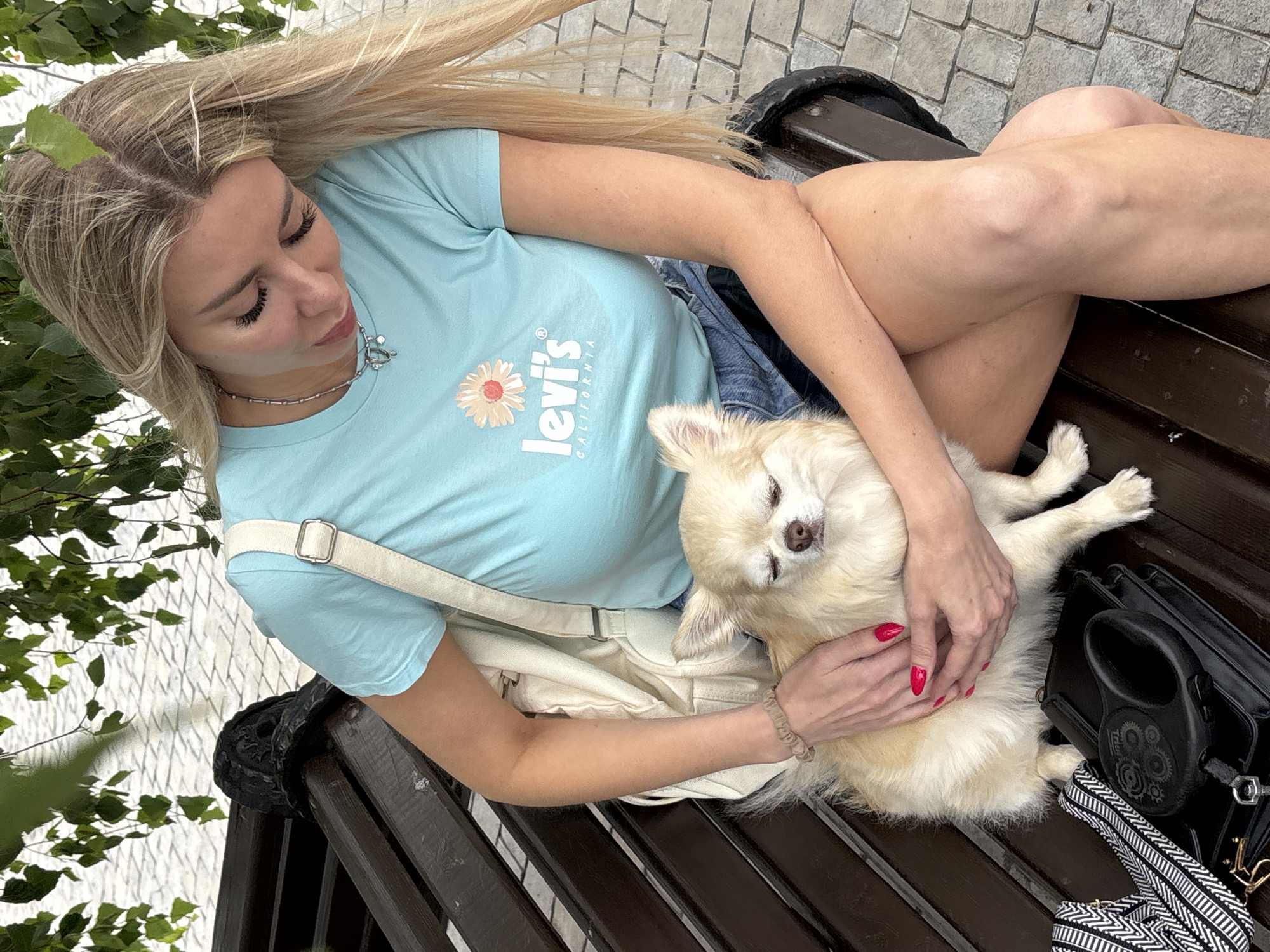 A blonde woman with long hair sits on a wooden bench under a tree with green leaves. She wears a light blue t-shirt with a Levi's logo and a daisy graphic, and denim shorts. The woman cradles a small, light-colored dog with its eyes closed in her lap. Her hands, with red nail polish, rest on the dog. A beige canvas bag with a white strap is slung over her shoulder. To her left on the bench, a black leather purse and a black retractable dog leash are placed. The background shows a cobblestone pathway and a glimpse of what appears to be a white building. The scene is consistent with a public park or outdoor seating area in Khryashchevka, Russia.