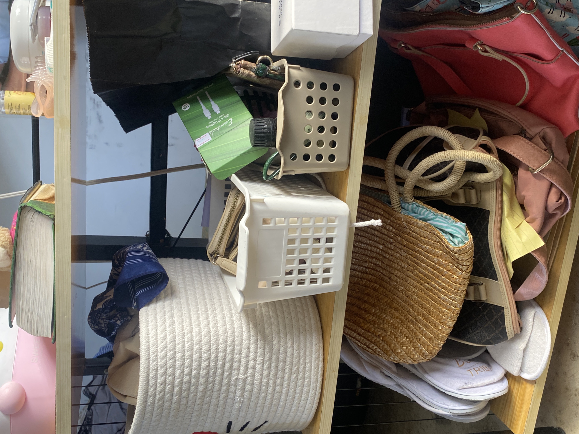The image shows a cluttered shelf inside a residential storage area in Davao City, Philippines. The shelf is made of wood and is divided into three tiers. The top tier displays a pink Hello Kitty container, a stack of books, and some assorted items. The middle tier contains a large white woven basket filled with dark fabric items, a small white plastic organizer with a coin purse, a beige plastic organizer with a small bottle and other items, and a white box. The bottom tier holds a brown woven bag, a black and beige handbag, a pink handbag, and several pairs of white slippers. The overall impression is one of disarray and the accumulation of personal belongings.
