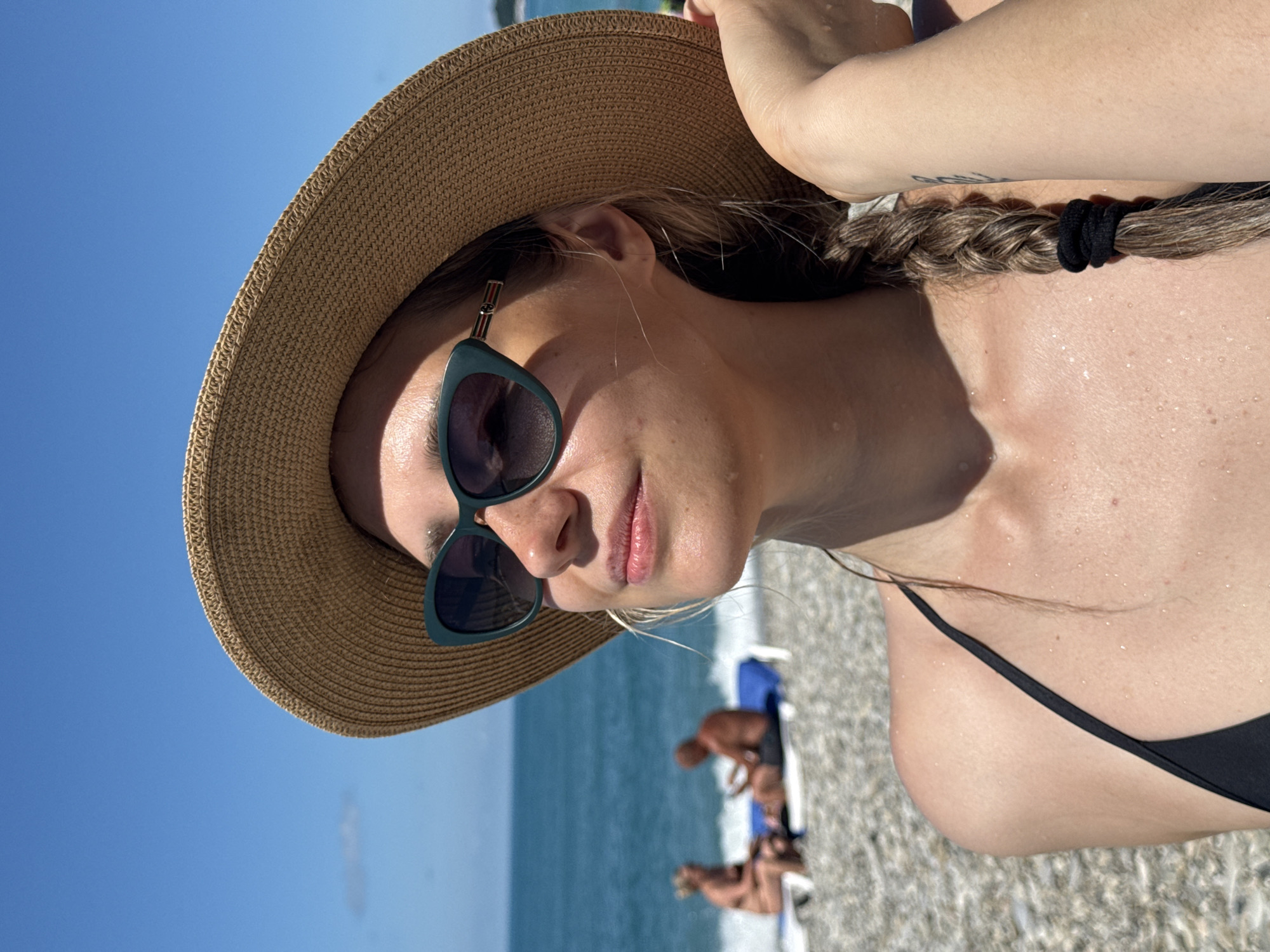 A woman with fair skin, a tan, and brown hair styled in a braid, is wearing a straw hat and dark sunglasses on a beach in Nebug, Russia. She is looking up and to the right, with a slight smile. Her shoulder and upper chest are visible, with water droplets on her skin, suggesting she has been in the water. She is wearing a black bikini top. In the background, two people are visible on lounge chairs near the ocean. The water is blue and calm, and the sky is clear. The beach is covered in pebbles.