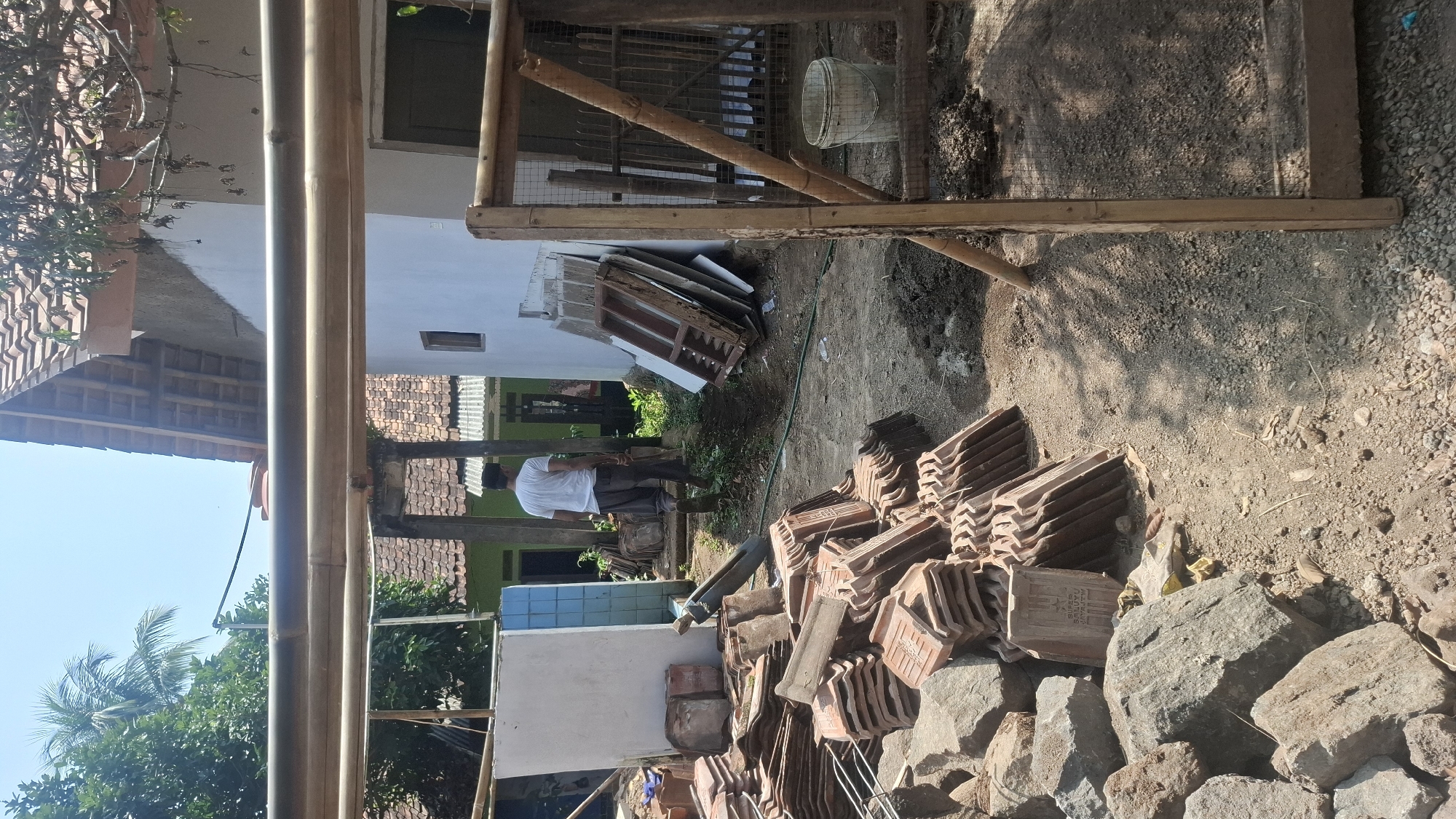 In Cibinong Hilir, Indonesia, an outdoor scene depicts a construction or renovation area. A man wearing a white t-shirt and dark pants stands in the middle ground, facing away from the camera, walking towards a white building. In the foreground and to the left, piles of terracotta roof tiles and large rocks are stacked on the dusty ground. To the right, a wooden frame structure, possibly part of a sieve or enclosure, is visible, with a white bucket placed inside. The background shows a white building with a tiled roof, and glimpses of other buildings and lush green trees, indicating a semi-rural or suburban environment under a clear blue sky.