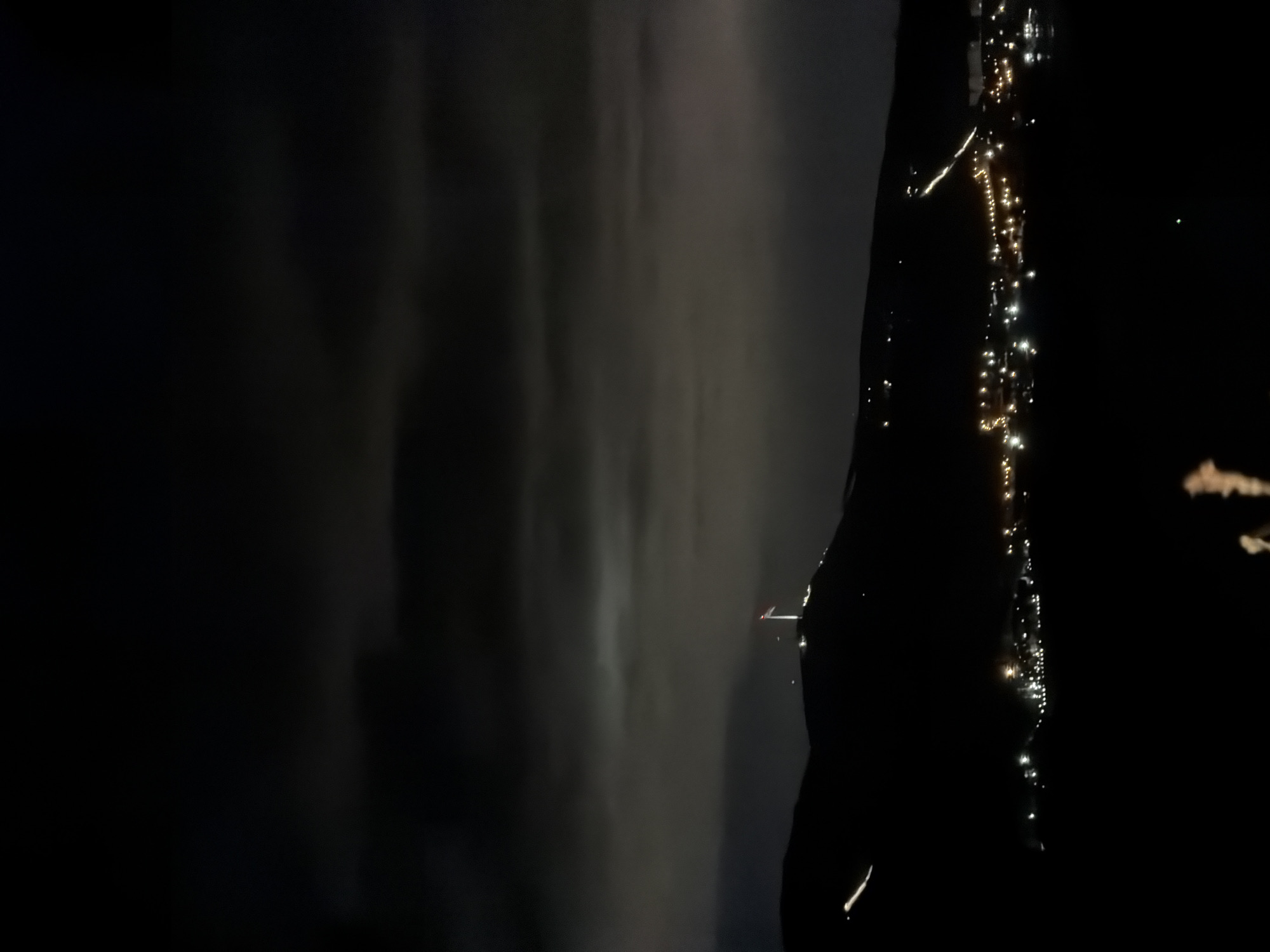 The image depicts a dark, night scene. On the right side, a dark, vertical figure stands out, possibly a person, against a backdrop of a line of yellow and white lights, likely indicating a cityscape or urban area. The lights appear to be scattered and may represent streetlights or illuminated buildings. To the left, there is a gray, hazy area that could represent fog, smoke, or poorly lit terrain. There are some glares or reflections visible. The image appears to be rotated ninety degrees. It is impossible to tell where in Слизнево, Russia this photo was taken, given the limited context.