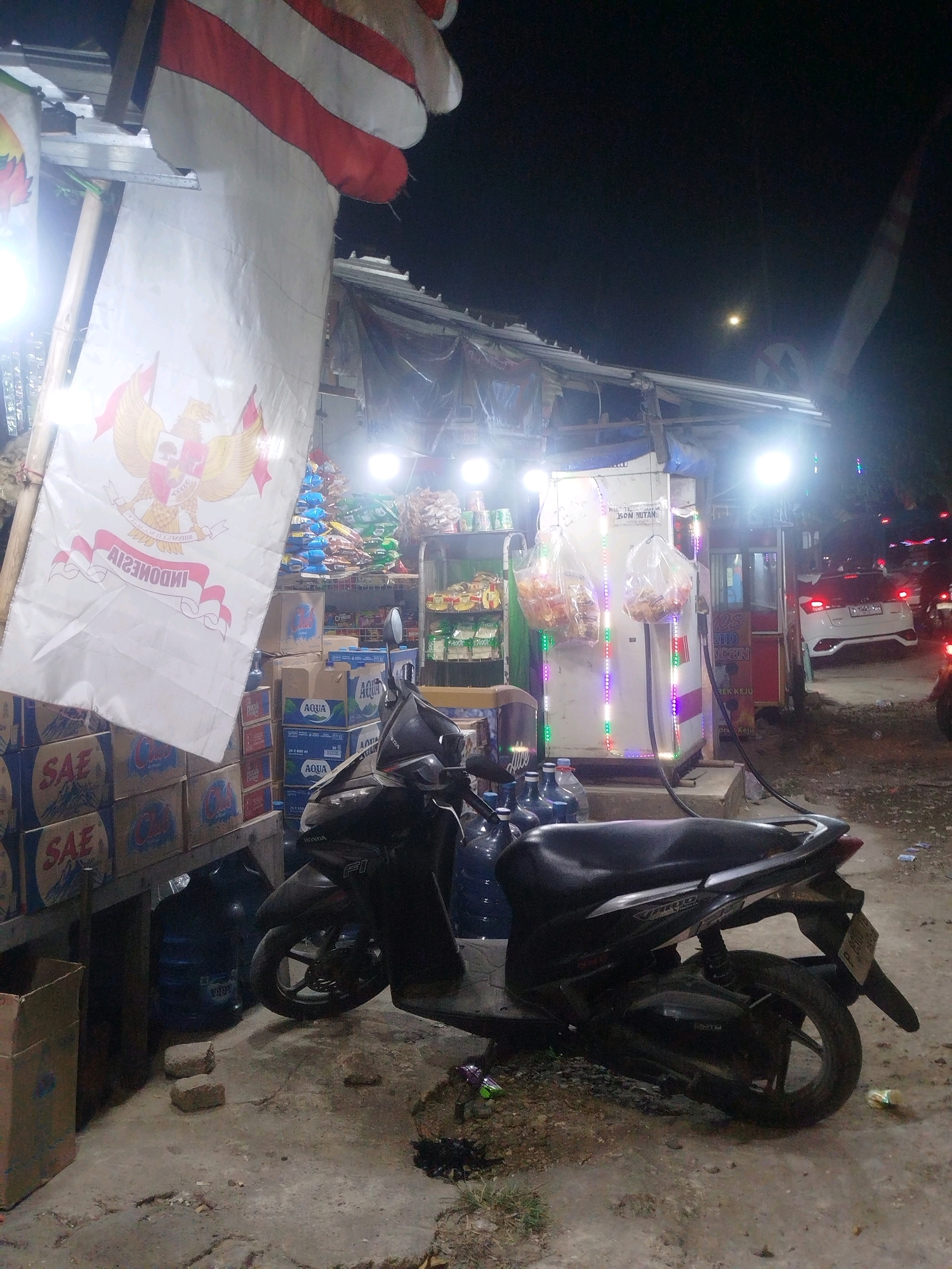 The image depicts a nighttime scene outside a small convenience store or roadside stall in Tonjung, Indonesia. In the foreground, a dark-colored scooter is parked on a paved surface. Behind the scooter, a stall is set up with shelves stocked with various goods, including bottled water and snacks. A white banner with the Indonesian coat of arms and text is prominently displayed on the left side of the stall. A fuel pump with colorful LED lights is visible in the center of the stall. In the background, a white car is parked, and the lights of other vehicles suggest a street or road nearby. The overall atmosphere is that of a bustling local business operating at night.