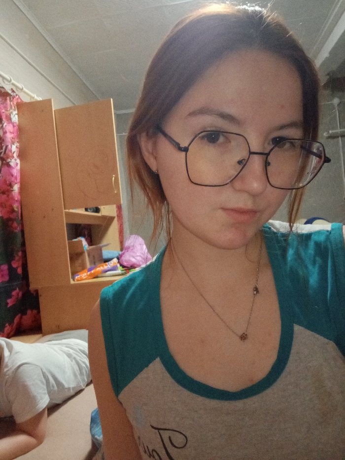 This image depicts an indoor scene in Старый Шагирт, Russia.
A young female, appearing to be in her late teens or early twenties, occupies the majority of the frame, facing directly towards the camera. She possesses light brown hair and wears black-framed, hexagonal-shaped eyeglasses. Her attire consists of a short-sleeved, two-toned top with a teal upper section and a light grey lower torso, featuring a partial dark inscription. A thin metallic necklace with two small, irregularly shaped charms is visible around her neck. Her expression is neutral.
In the lower-left quadrant, a second individual is partially visible from the back, wearing a white short-sleeved t-shirt. Only their upper back and right arm/elbow are discernible, indicating a prone or kneeling position on a light-colored surface.
The background reveals a domestic setting. To the left, a light-colored wooden furniture unit, possibly a shelf or wardrobe, is present, with one door slightly ajar and various items visible within its compartments, including a crumpled purple textile item, an orange and white cylindrical object, and a cardboard box. Adjacent to the furniture, a curtain adorned with a pattern of pink and red flowers on a dark background hangs from a light-colored rod. The ceiling is constructed from light-colored panels.