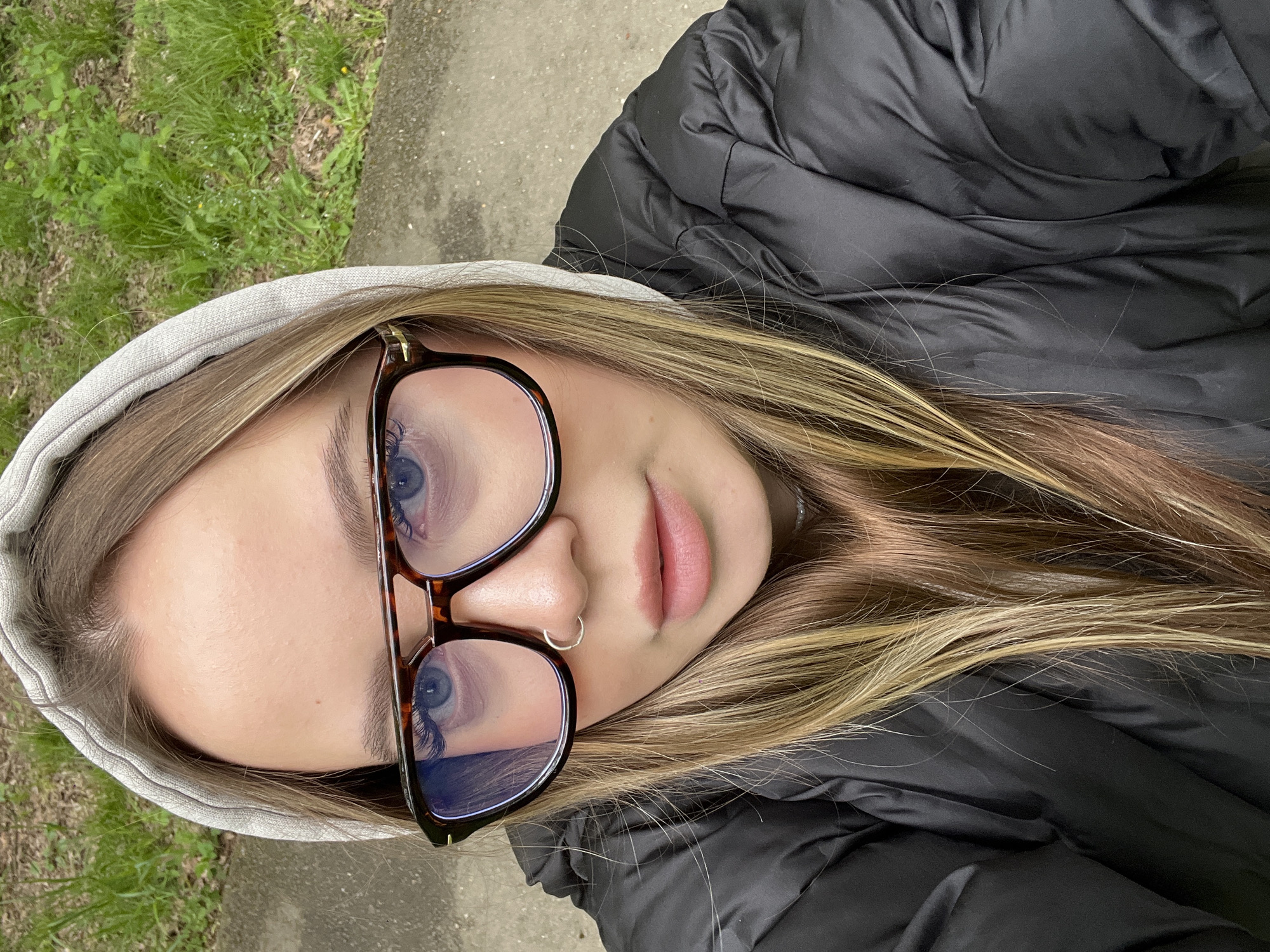 A close-up, top-down selfie features a young person, likely female, captured outdoors in what appears to be Krasnodar, Russia. The subject's face is prominent, showcasing blue eyes framed by eyeglasses with dark, possibly tortoiseshell, frames. Their long, light brown hair with blonde highlights spills around their head and shoulders. A small silver hoop nose ring adorns the right nostril.
The person is wearing a light grey hooded sweatshirt, with the hood pulled up around their head, and a black puffer jacket, visible over their shoulders and upper body. The jacket has a slightly shiny, wrinkled texture.
The background suggests an outdoor setting. To the left and upper part of the image, green grass with some weeds and lighter patches is visible. A grey concrete or paved surface runs along the right and lower edges, and also appears beneath the grass area. The perspective indicates the photo was taken from above, with the subject looking upwards towards the camera.
The lighting suggests it is daytime. The subject has a neutral to slightly relaxed expression, with their lips slightly parted. There is no visible text within the image.